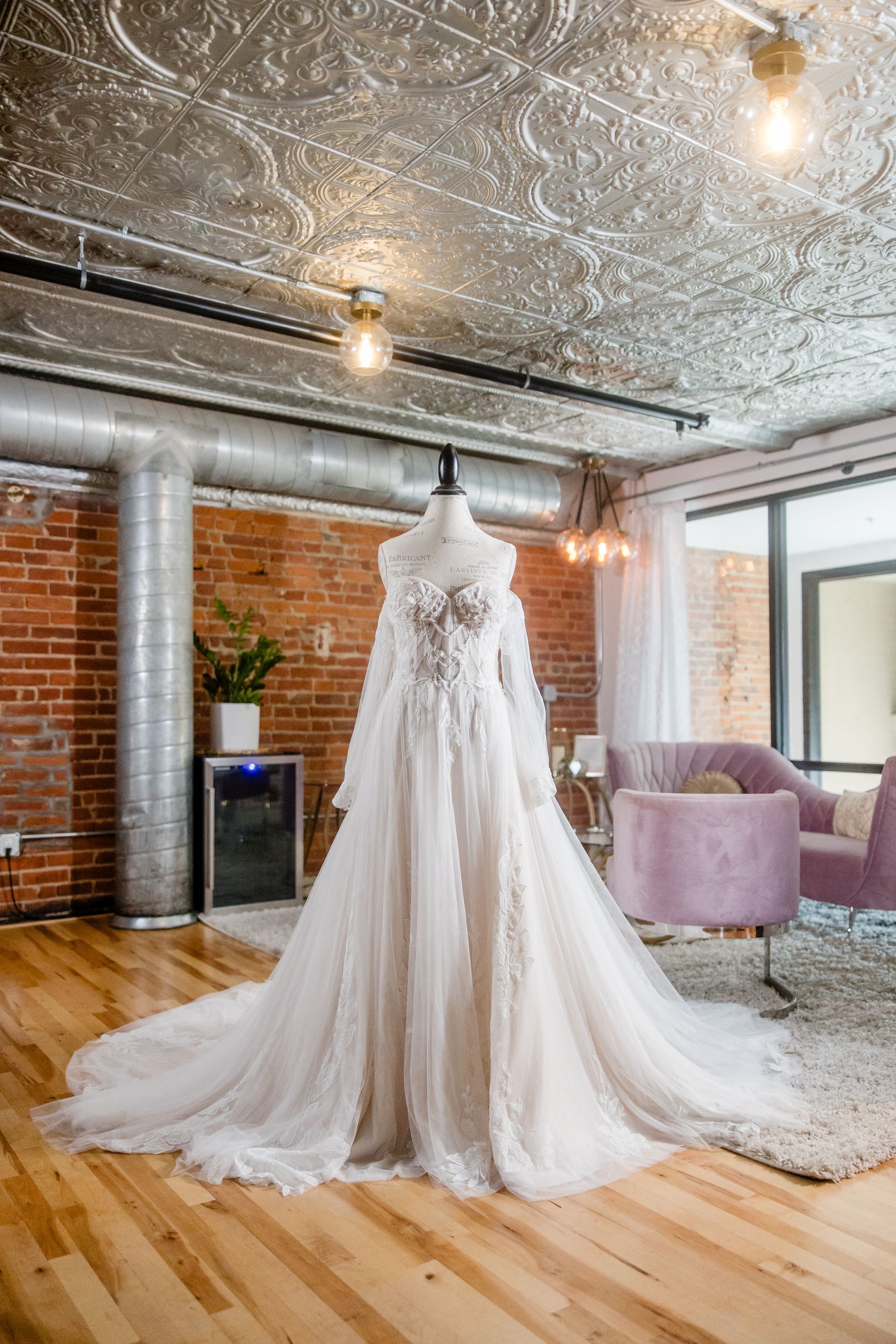 White wedding dress on mannequin in a brick-and-wood studio with ornate ceiling and purple chair