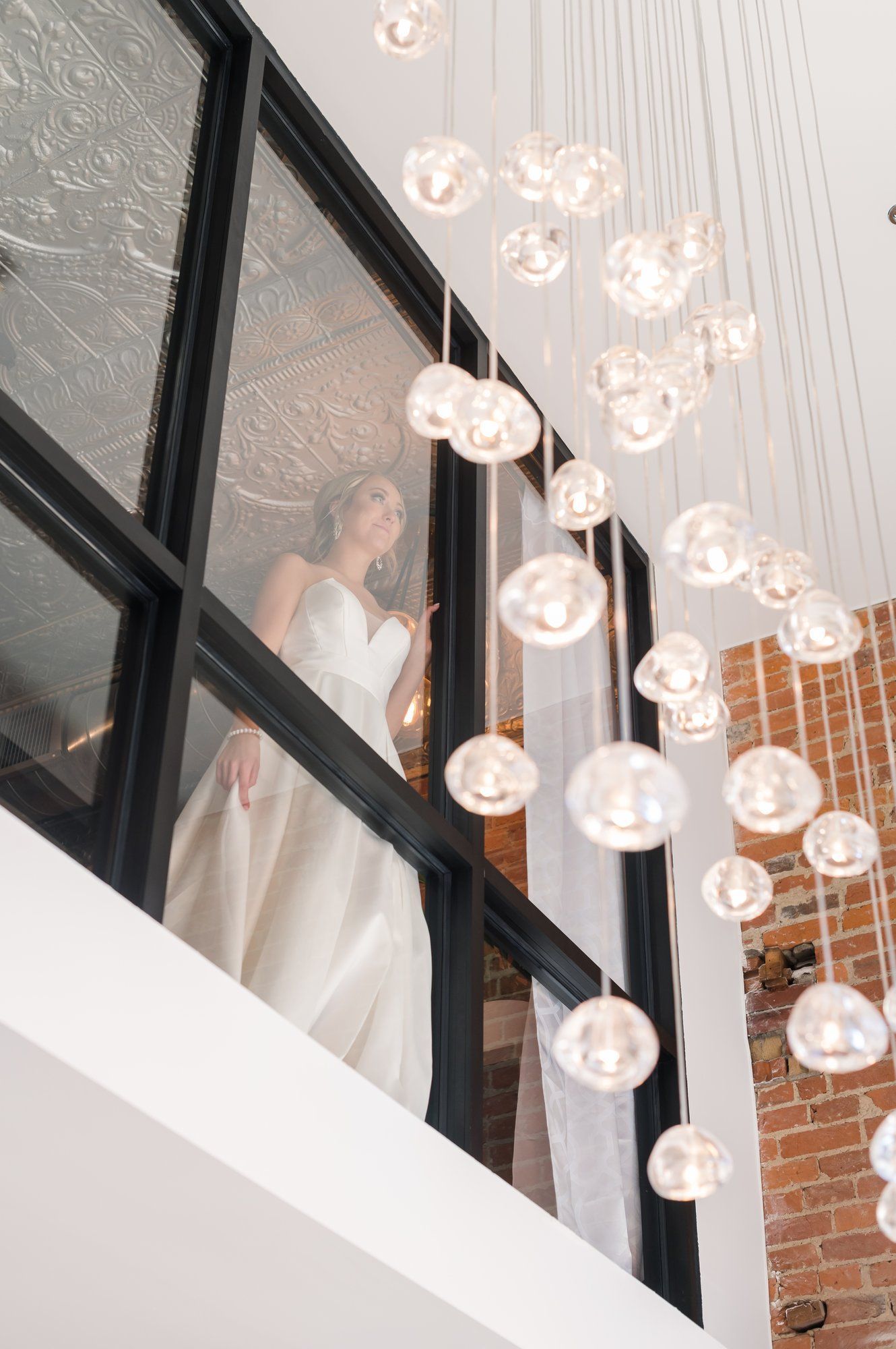 Bride in a white gown standing by a black-framed window beside hanging glass sphere lights