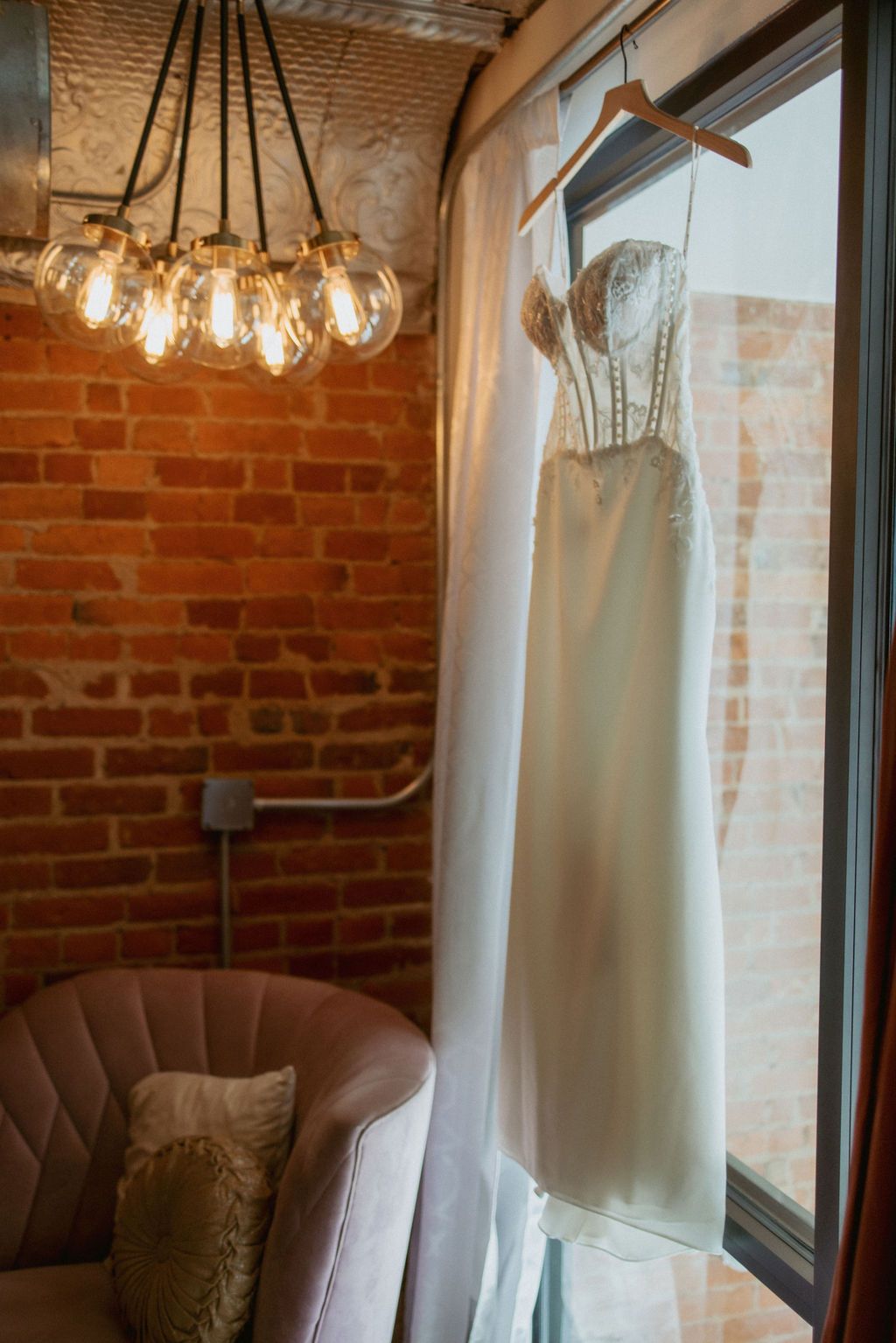 White dress hanging by a window in a cozy brick-walled room with a chandelier and chair