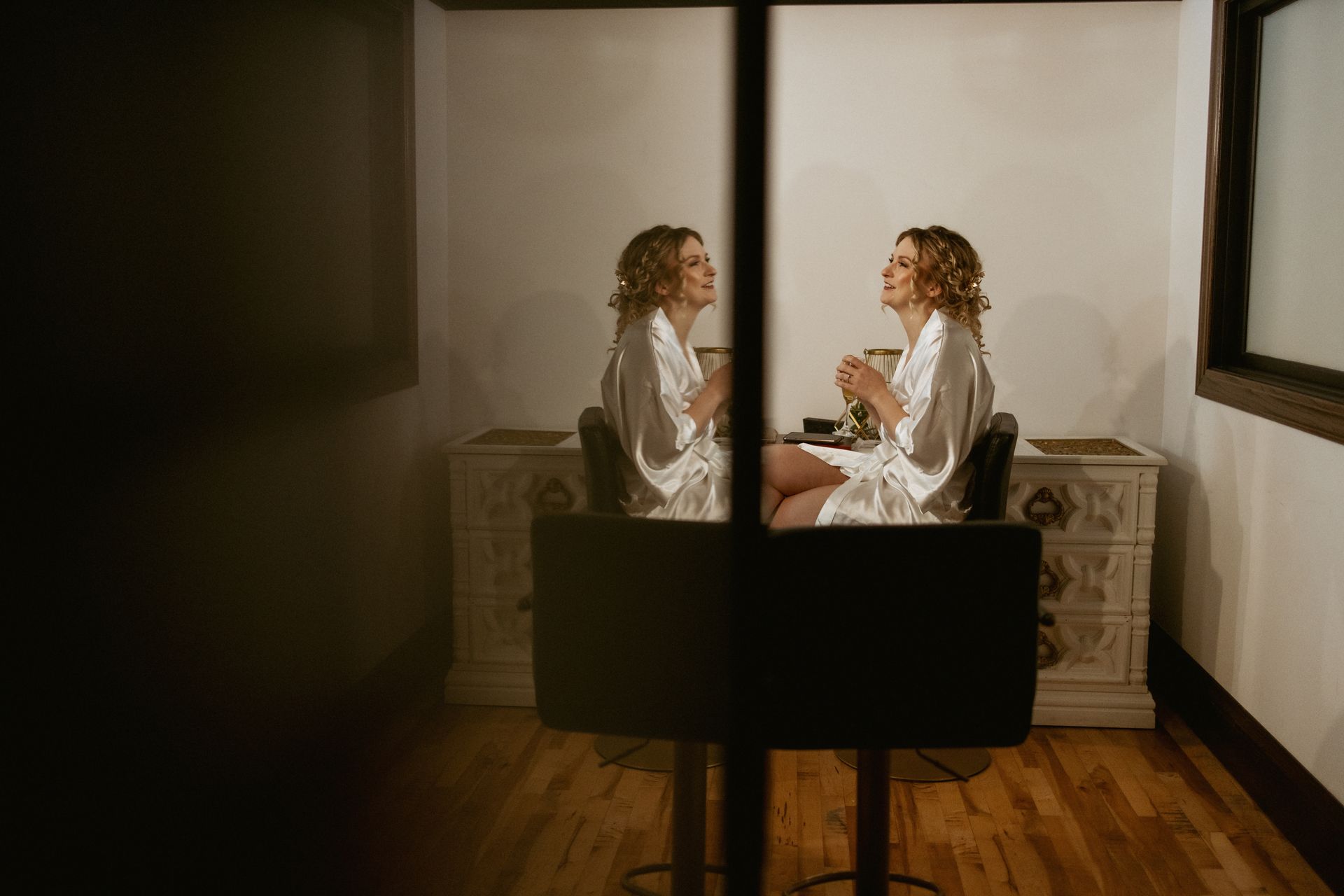 Woman seated at a vanity, looking into a mirror in a softly lit room.