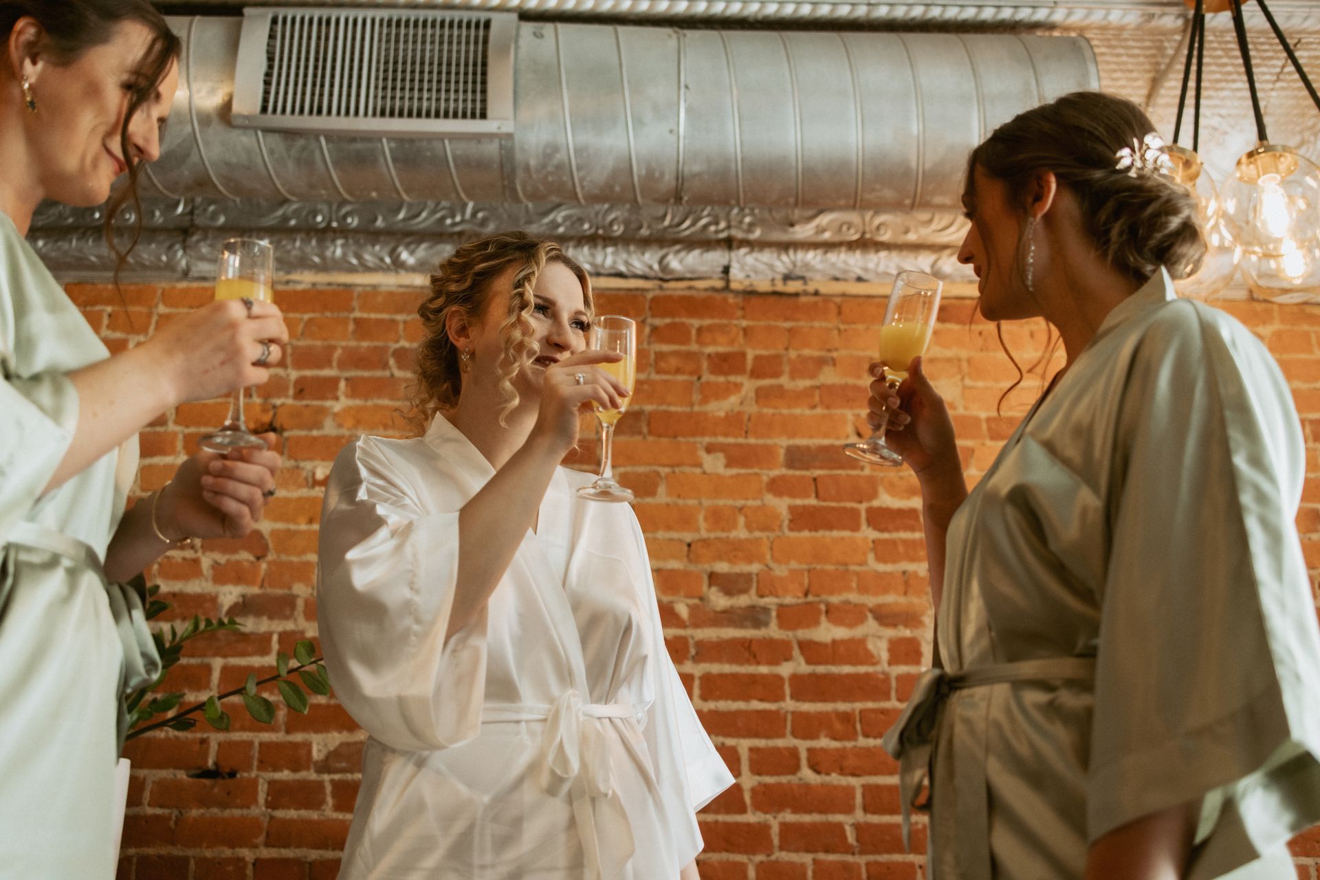 Three people in white robes raise drinks in a loft with brick walls and hanging glasses.