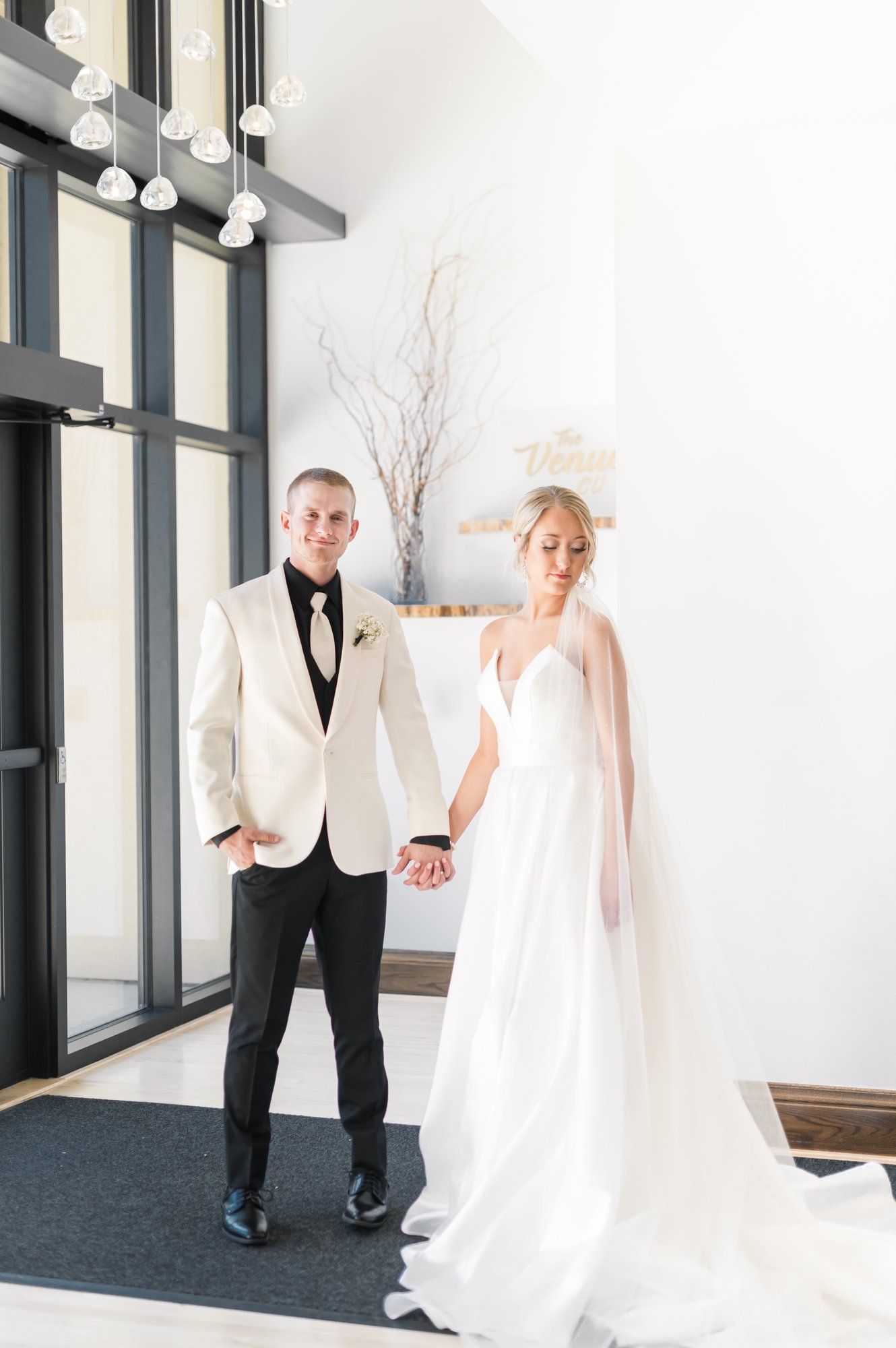Bride and groom holding hands in a bright indoor venue, wearing a white gown and cream jacket