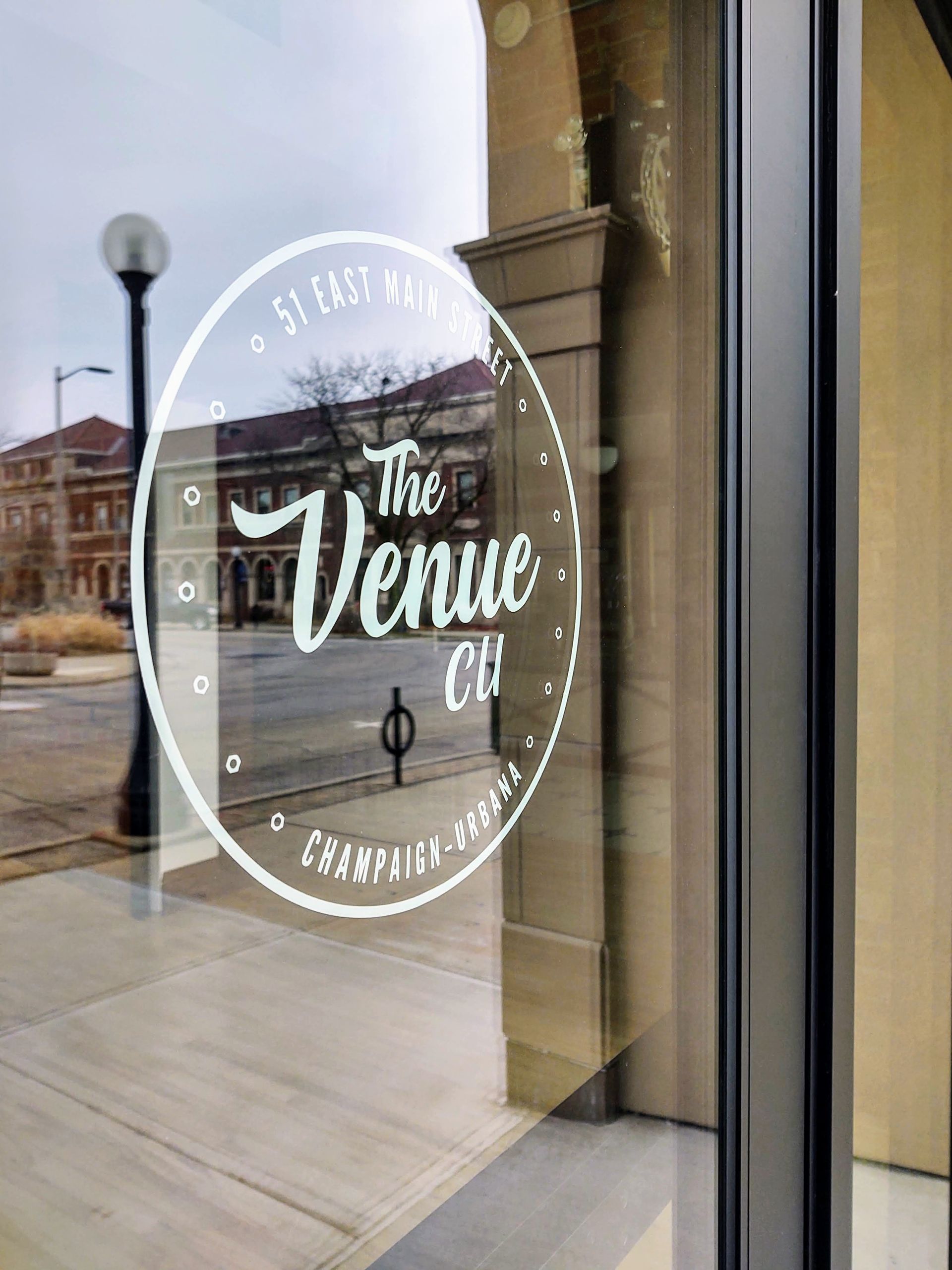 White oval storefront decal reading “The Venue” on a glass door with a street reflected outside