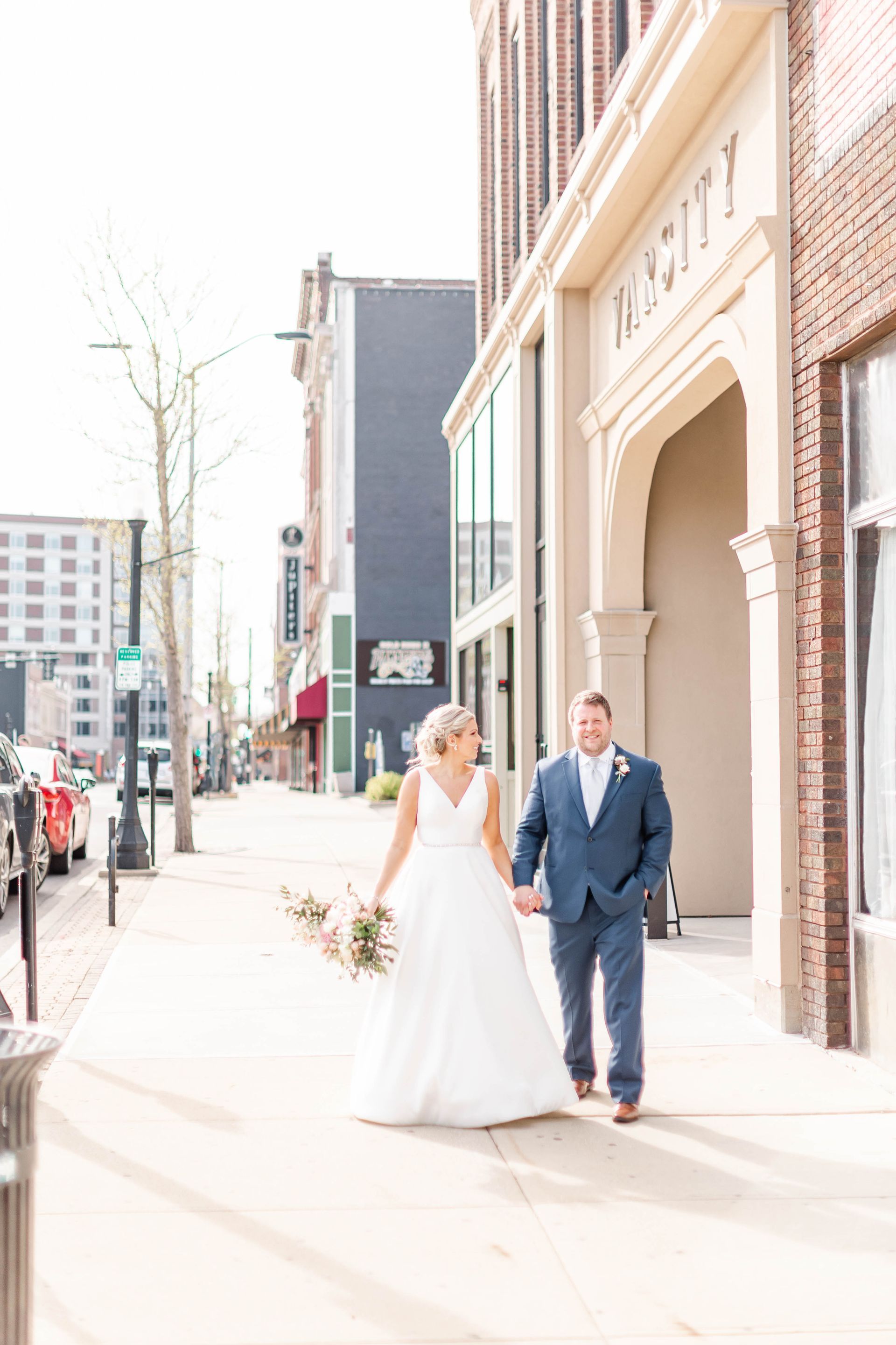 Bride and groom walking on a sunny city sidewalk beside brick buildings