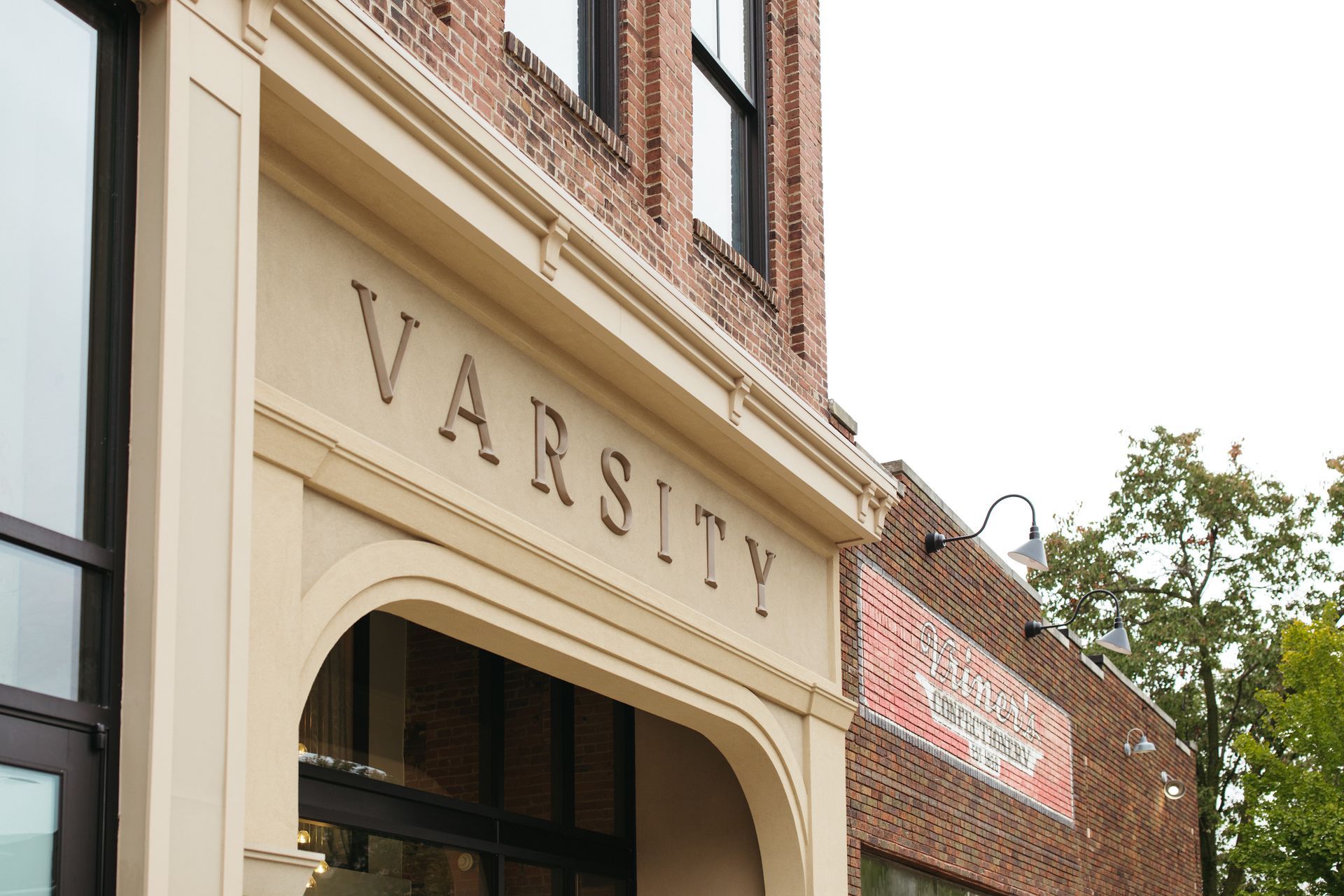 Storefront with “VARSITY” sign on a cream building beside brick walls and trees