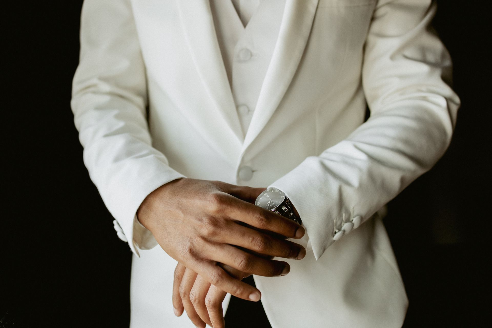 Person in a white suit adjusting a wristwatch against a dark background