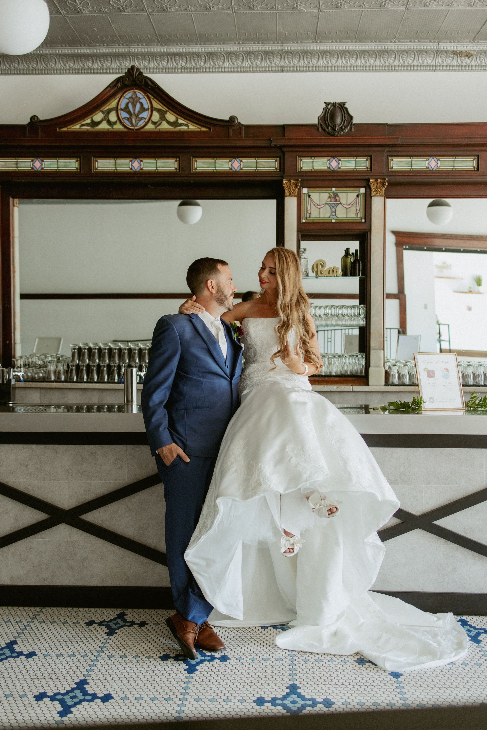 Couple in wedding attire kissing at a bar with ornate wood trim and blue tile floor