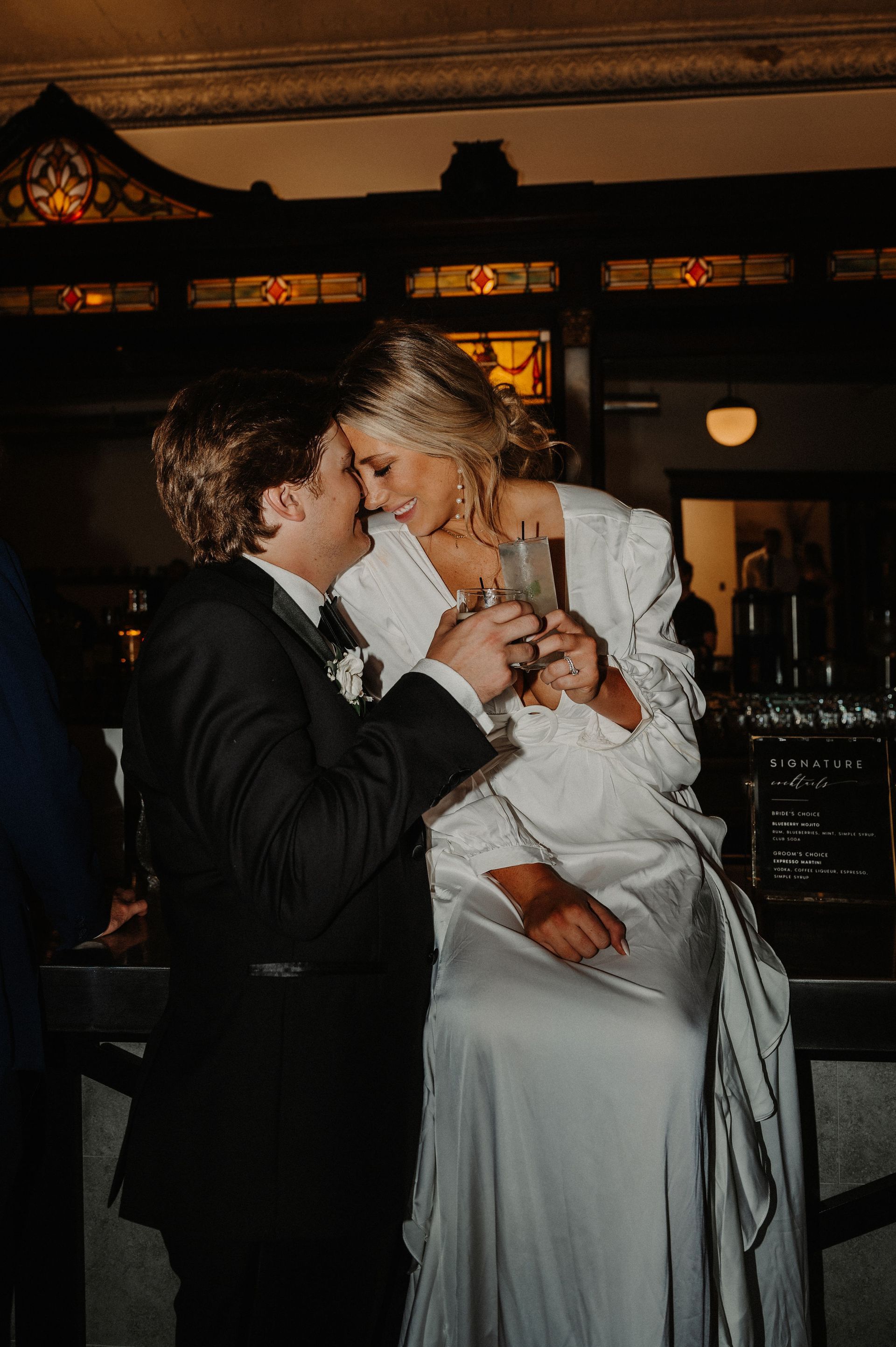 Two people in formalwear kissing while seated together in a warmly lit bar or lounge