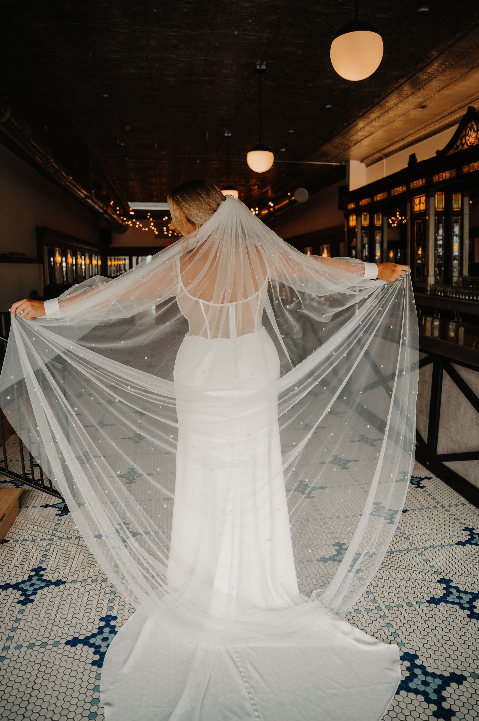 Bride in a white gown with a long veil, standing in a dimly lit corridor with hanging lanterns.