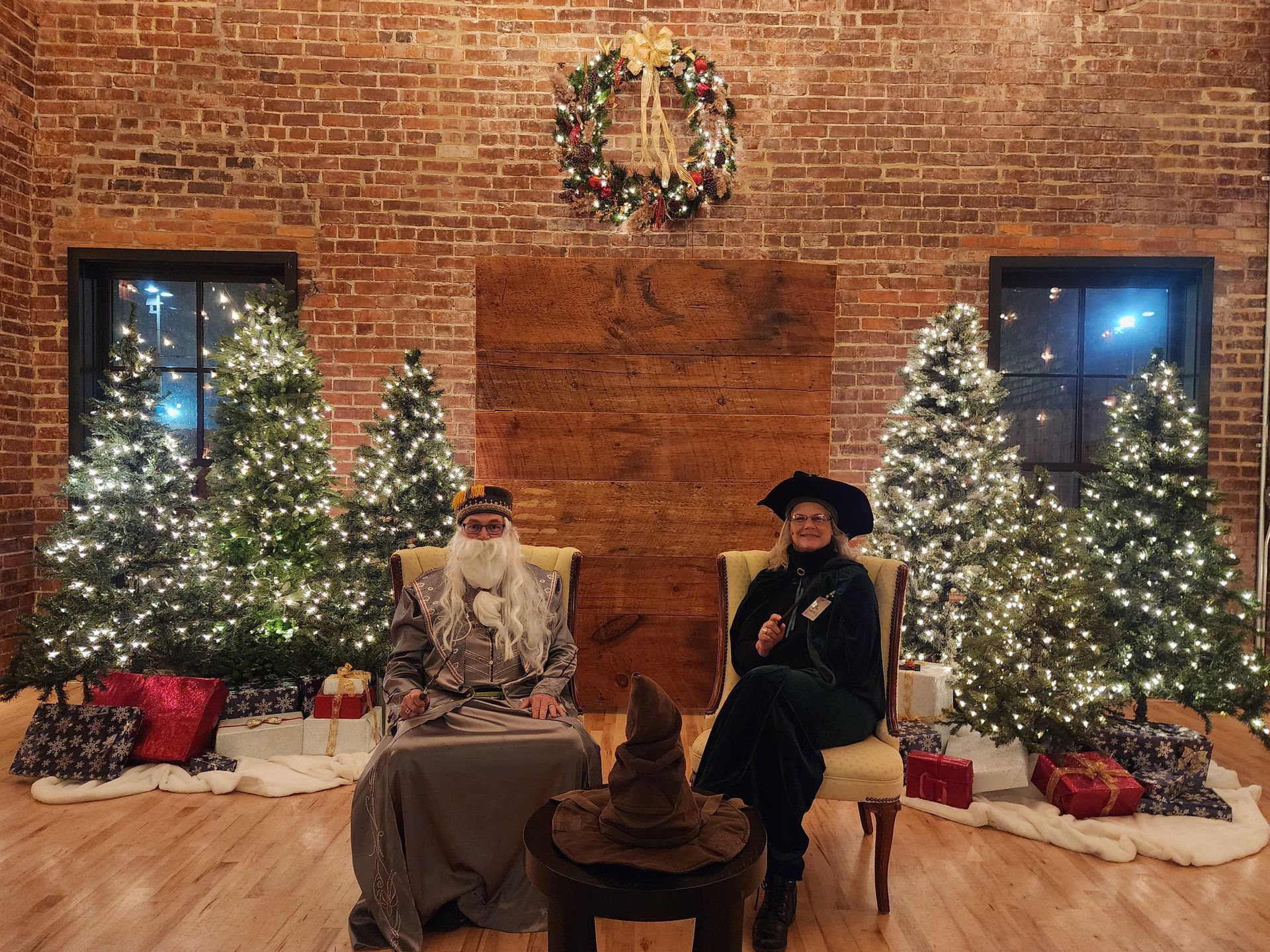 Two people seated by lit Christmas trees and gifts in front of a brick fireplace wall