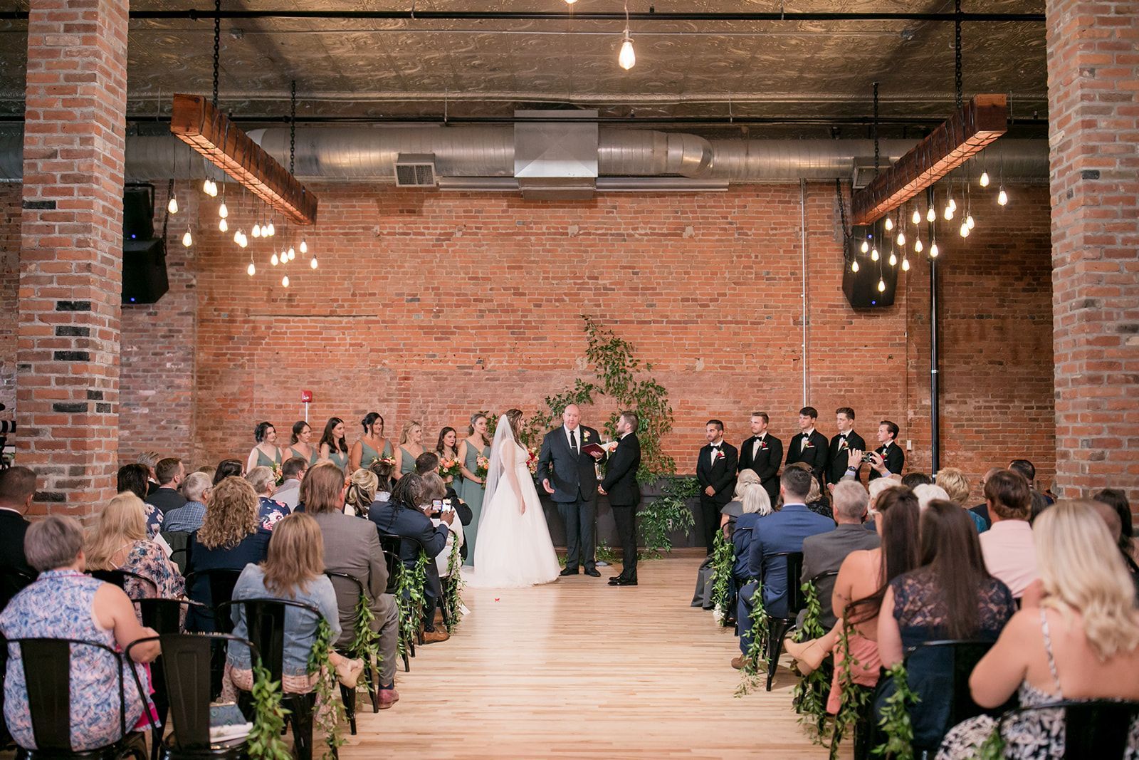 Wedding ceremony in a brick-walled hall, bride and groom at the altar with guests seated on both sides.