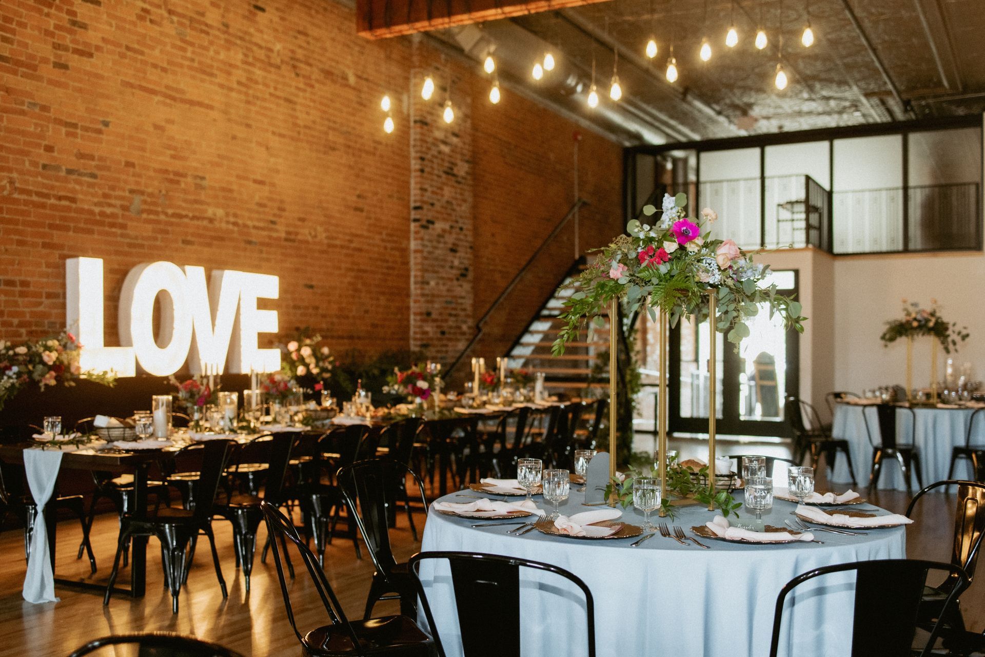 Elegant wedding reception in a brick loft with a glowing “LOVE” sign, long tables, and floral centerpieces