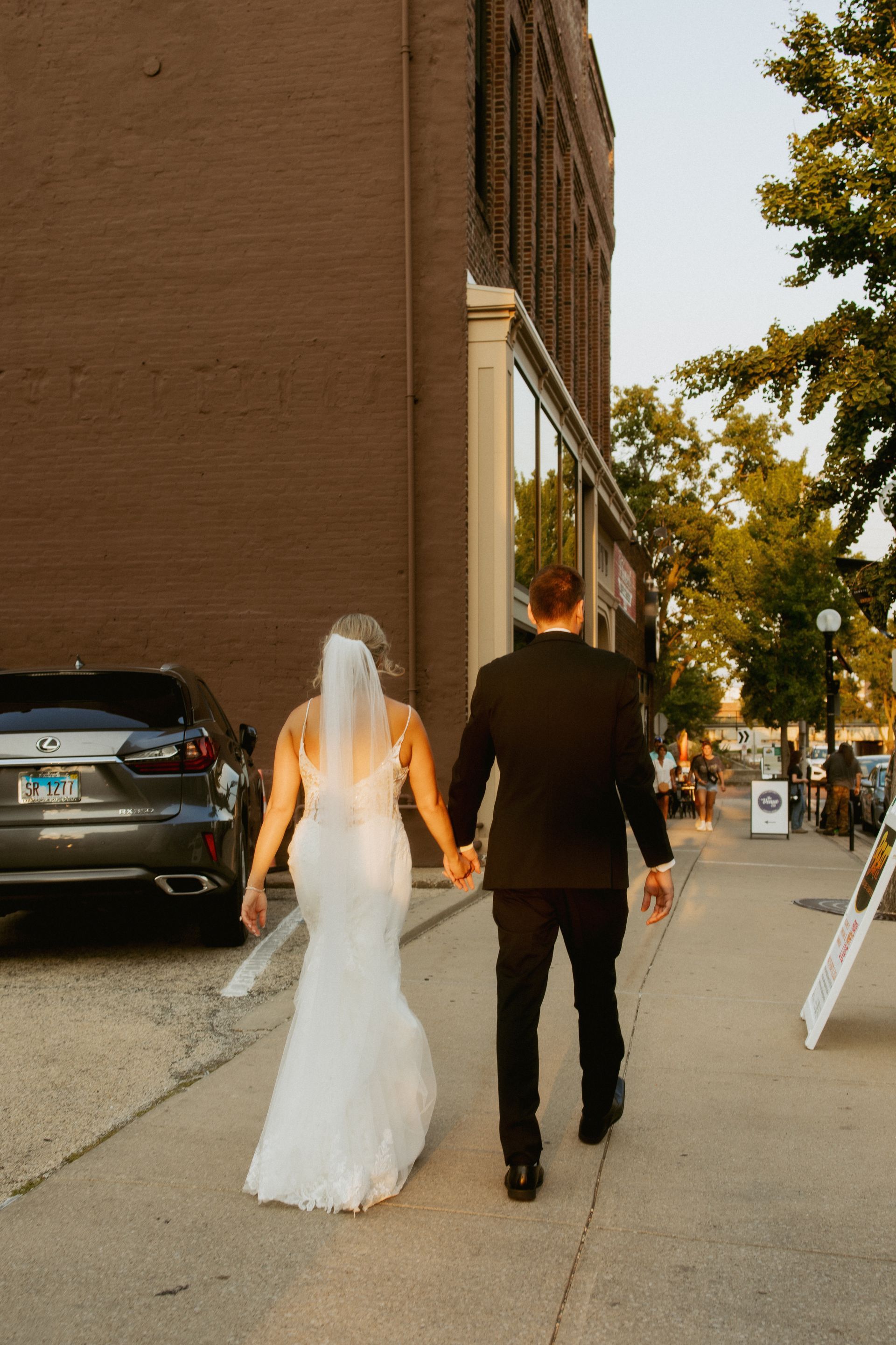 Bride and groom in wedding attire walking down a sidewalk beside a brick building