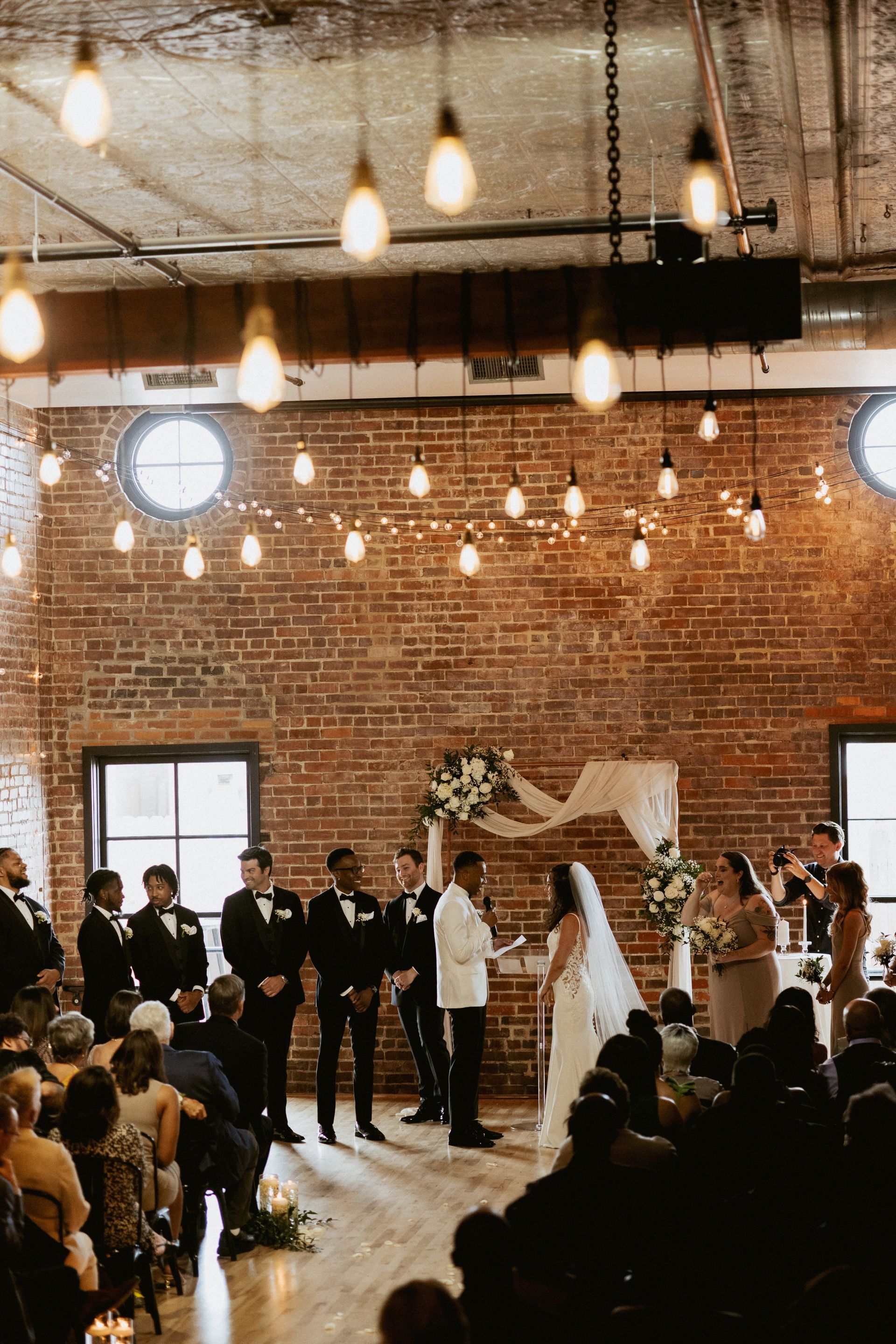 Wedding ceremony in a brick loft venue with hanging lights, bride, groom, officiant, and seated guests.