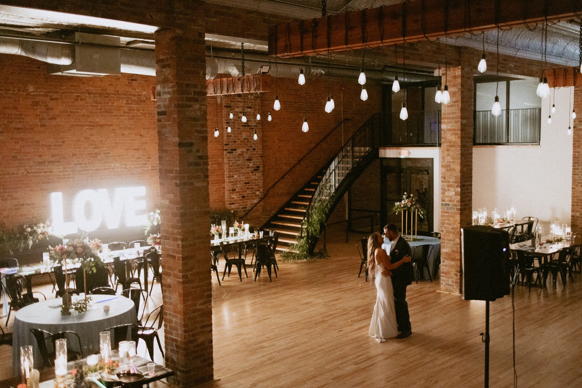 Wedding reception in a brick loft venue, with a glowing “LOVE” sign and a couple dancing on the wooden floor