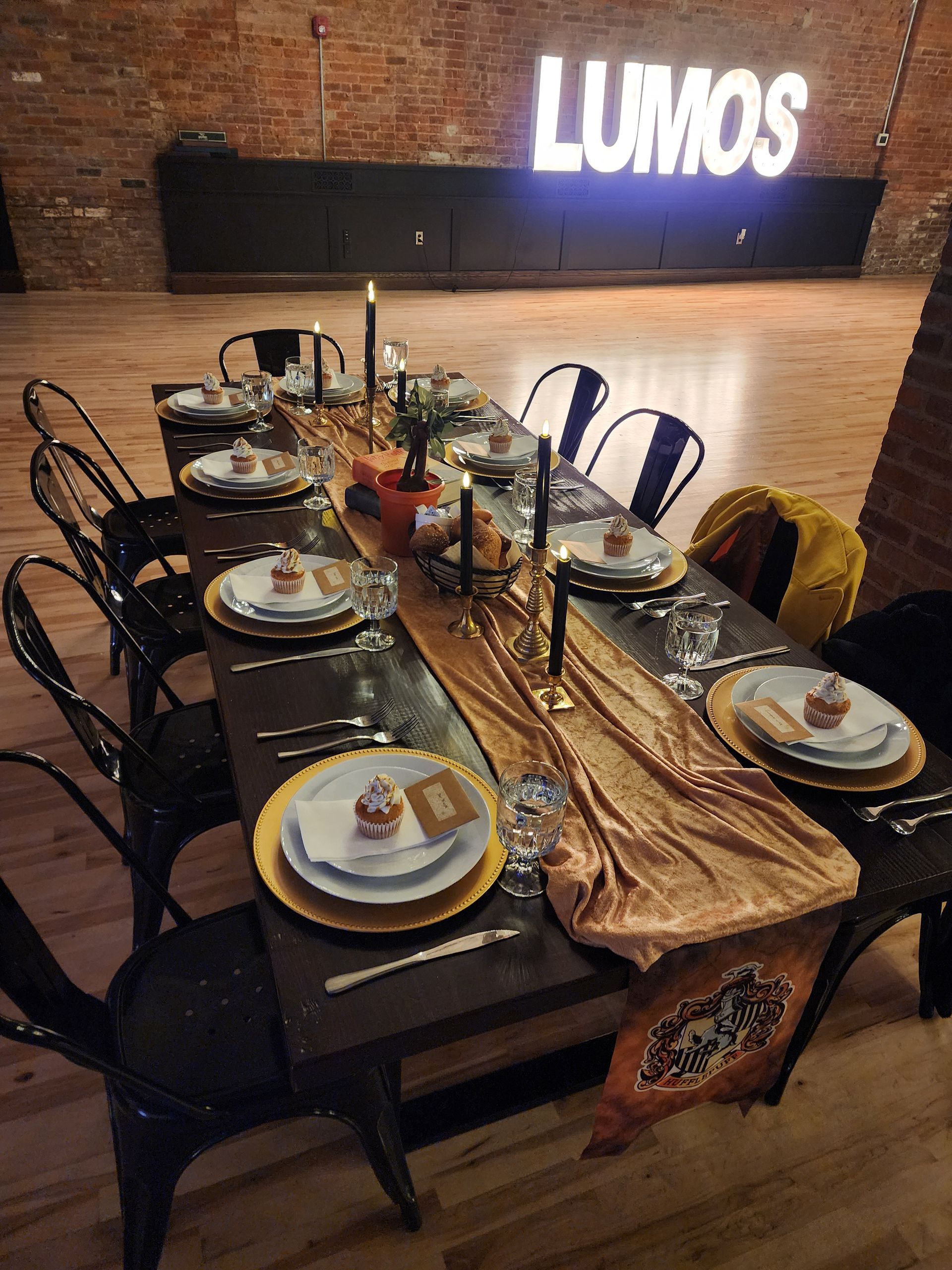 Long dining table set with plates and glasses in a dim event space, with glowing “LUMOS” sign behind it