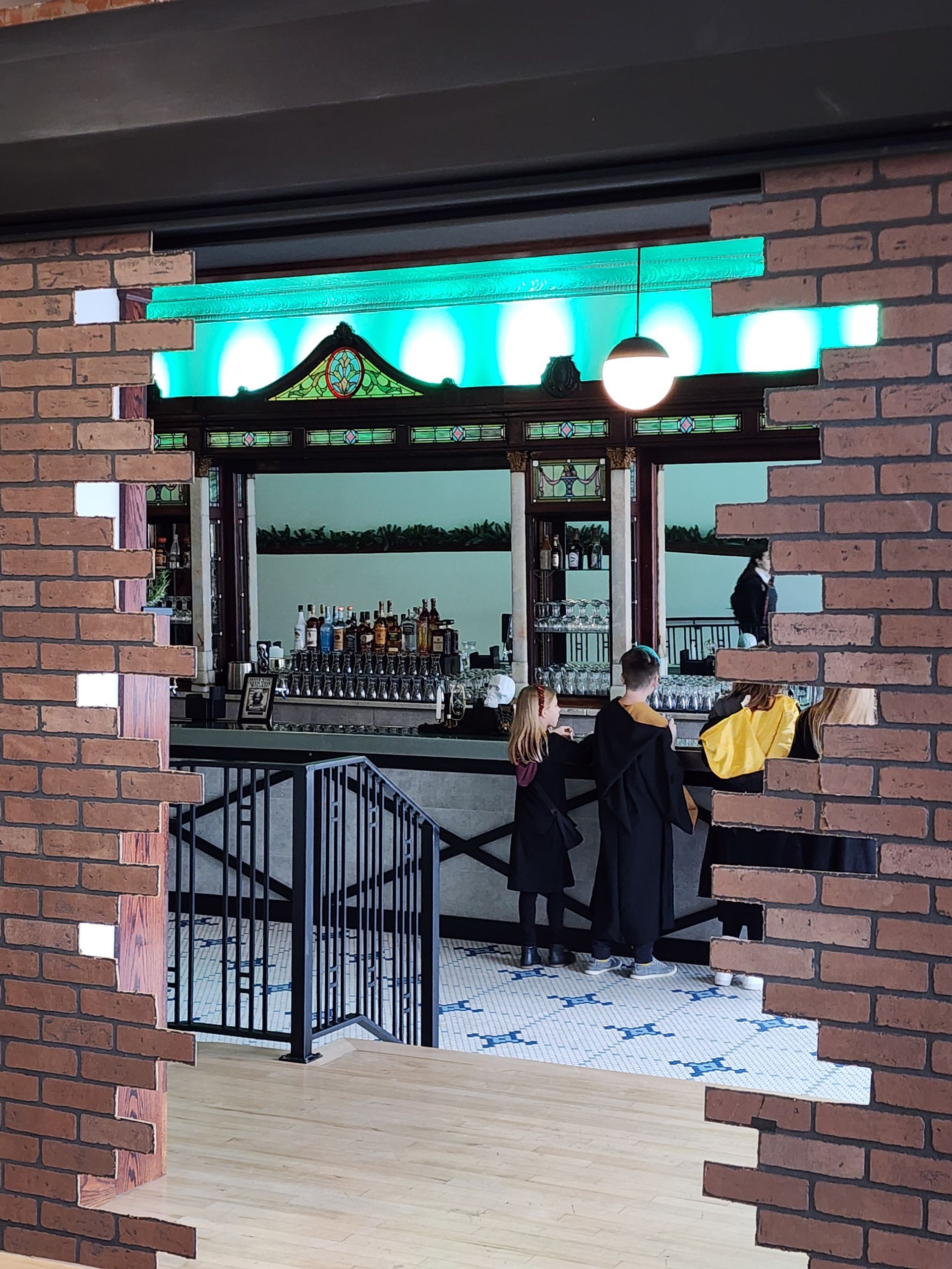 People standing at a bar seen through a brick wall opening, with teal neon lights and hanging lamps