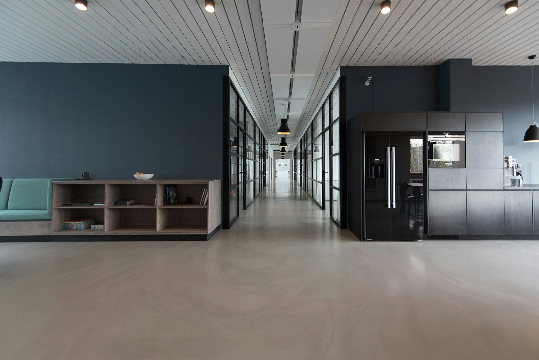 Modern open office with dark work pods, kitchen area, and a long central corridor beneath ceiling lights