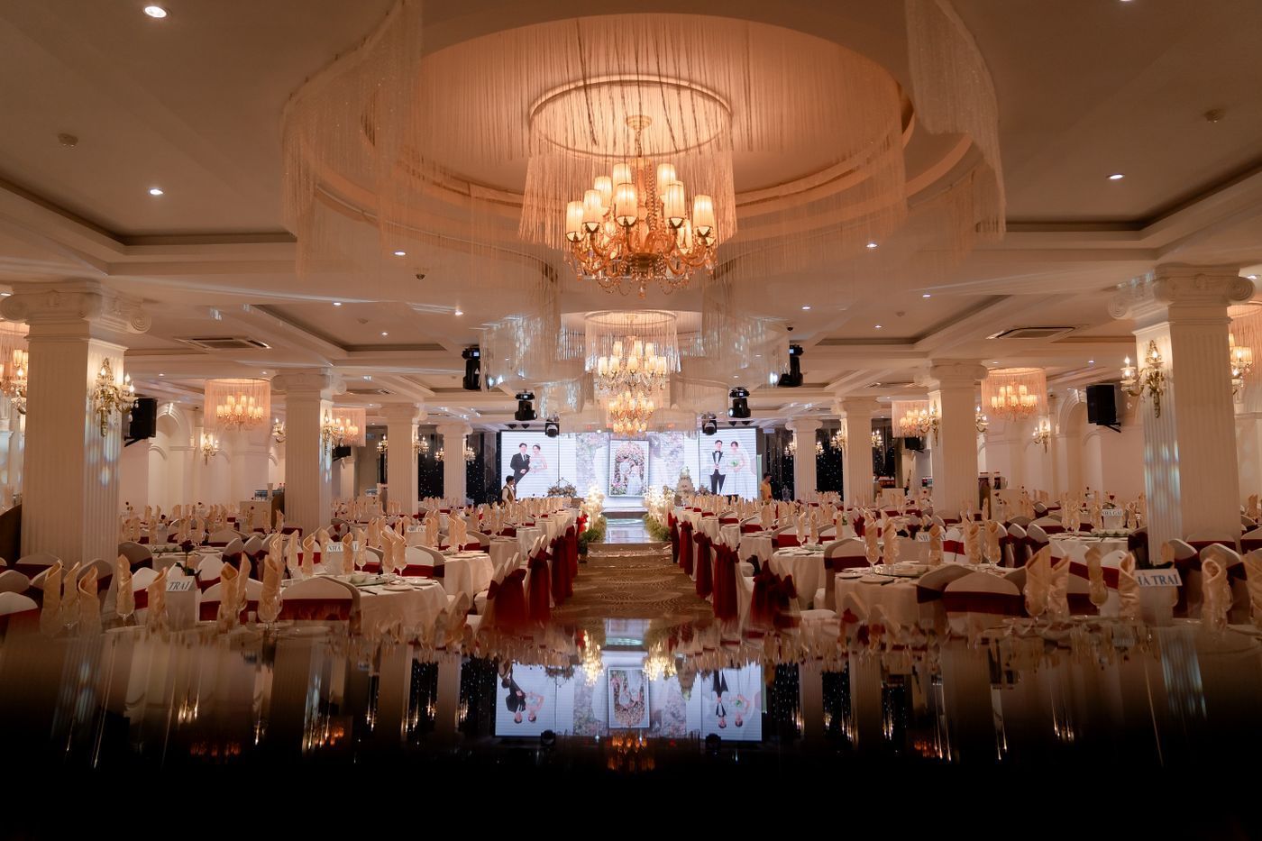 Elegant banquet hall with chandeliers, round tables, and white chairs reflected on a polished floor.