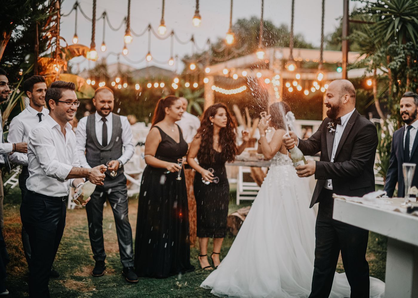 Wedding guests toast the bride and groom under string lights at an outdoor evening reception.