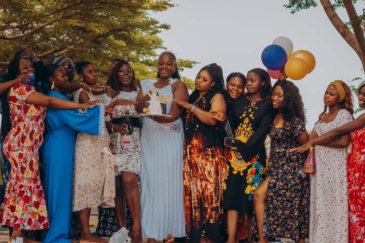 Group of people celebrating outdoors, gathered around a cake with colorful balloons and trees in the background.