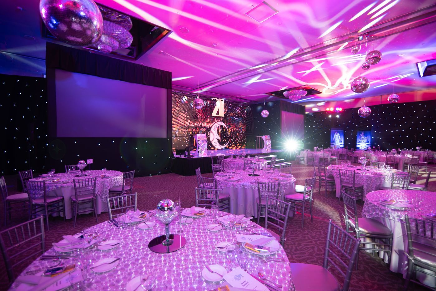 Lavishly decorated banquet hall with round tables, pink-purple lighting, and a large projection screen