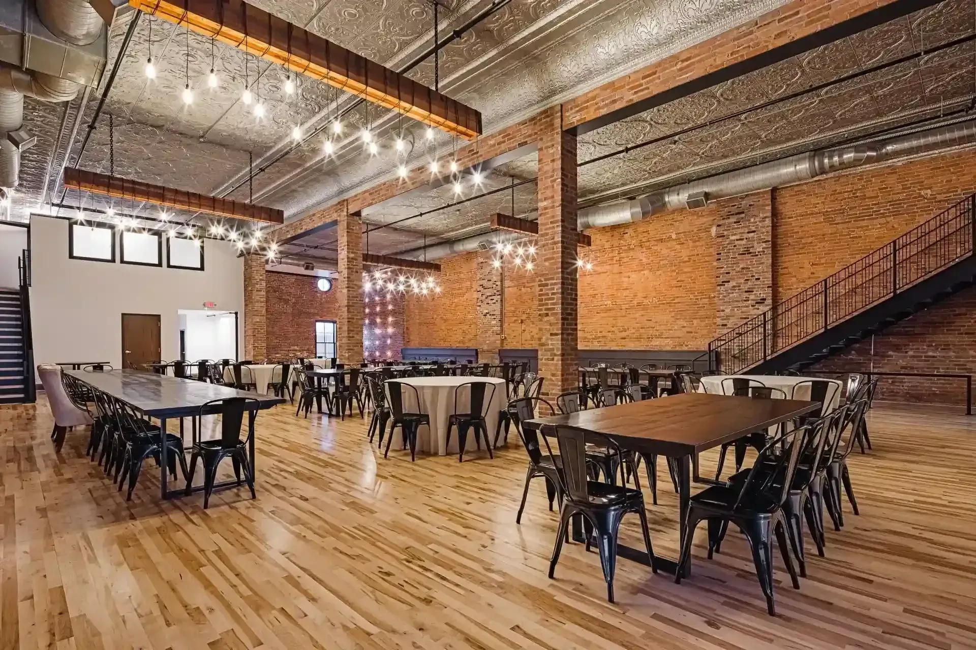 Spacious industrial-style dining hall with wooden floors, brick walls, tables, and chairs under string lights