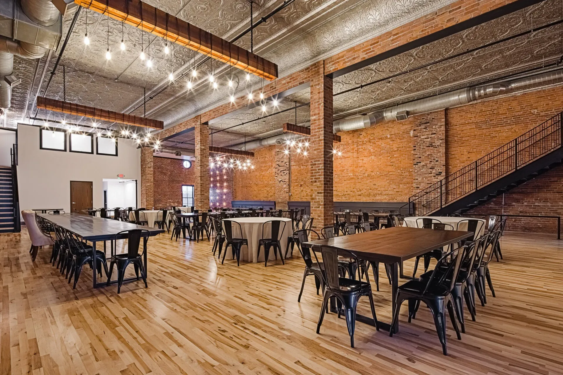 Spacious industrial-style dining hall with wooden floors, brick walls, tables, and chairs under string lights