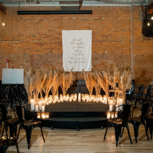 Decorative wedding altar with candles, pampas grass, and black chairs against a brick wall