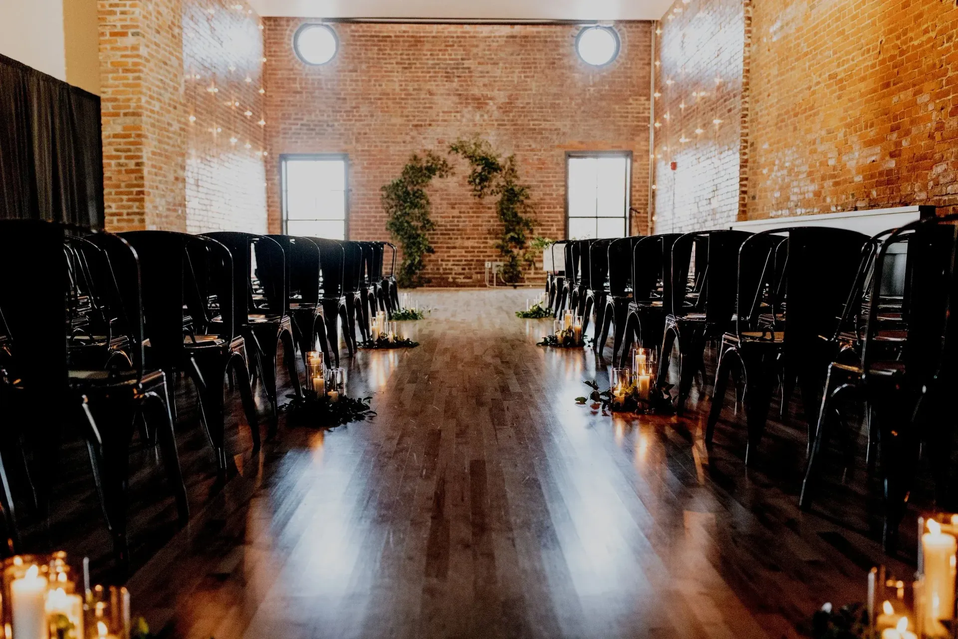 Empty banquet hall with rows of black chairs, warm brick walls, and candlelit aisle reflecting on polished wood floor