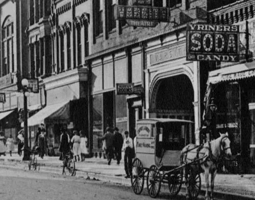 Black-and-white street scene with pedestrians, storefronts, and a horse-drawn carriage