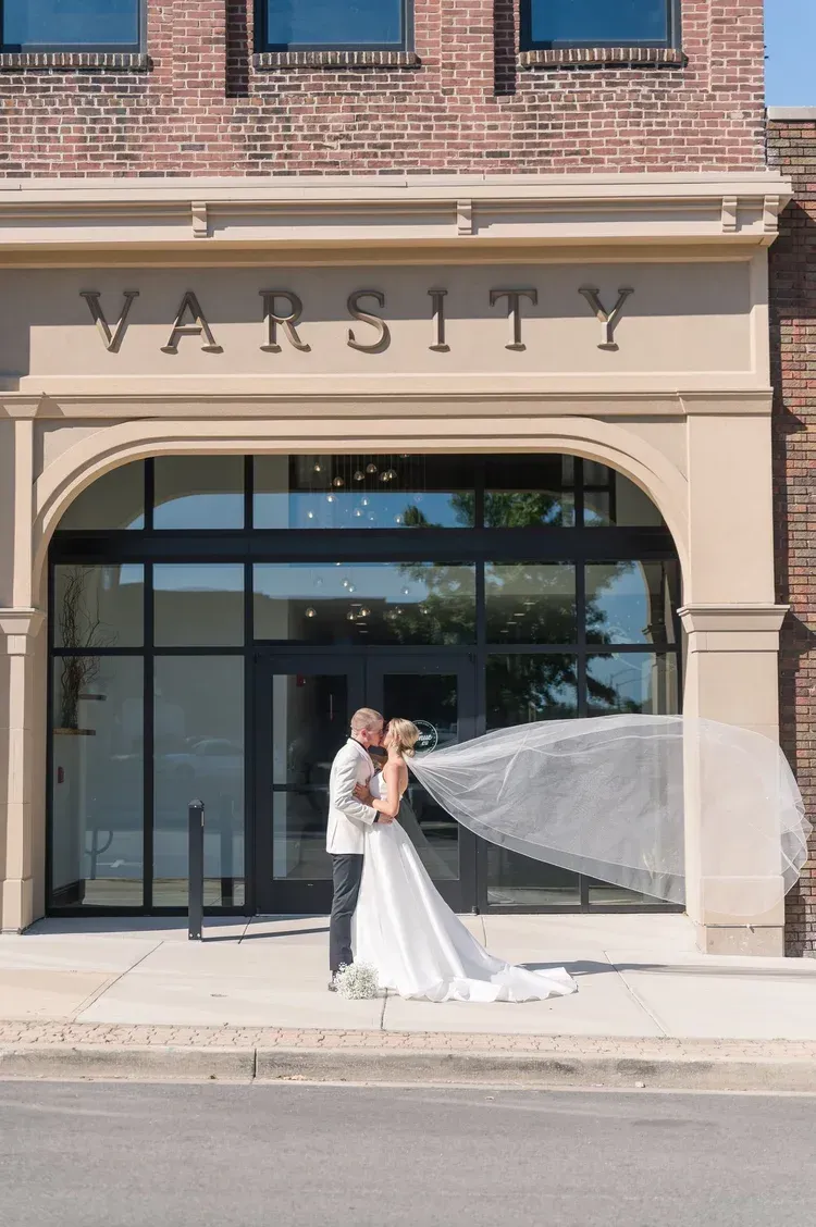 Bride and groom posing outside Varsity storefront, veil blowing in the wind