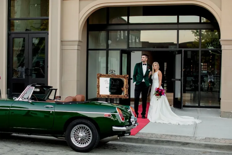 Bride and groom stand outside a building beside a green vintage car with a red carpet.