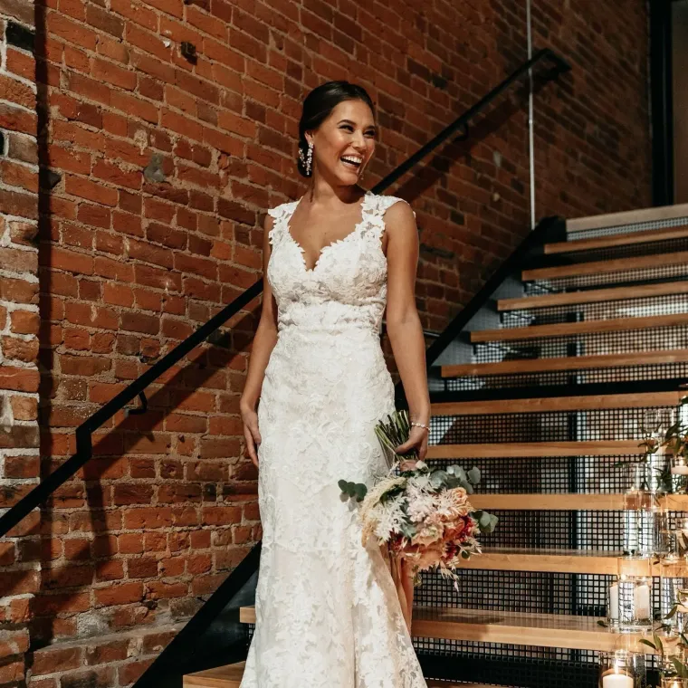 Bride in a lace white gown holding a bouquet on a staircase against a brick wall
