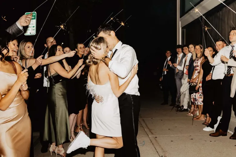 Couple kissing at a night wedding send-off, surrounded by guests cheering and holding sparklers