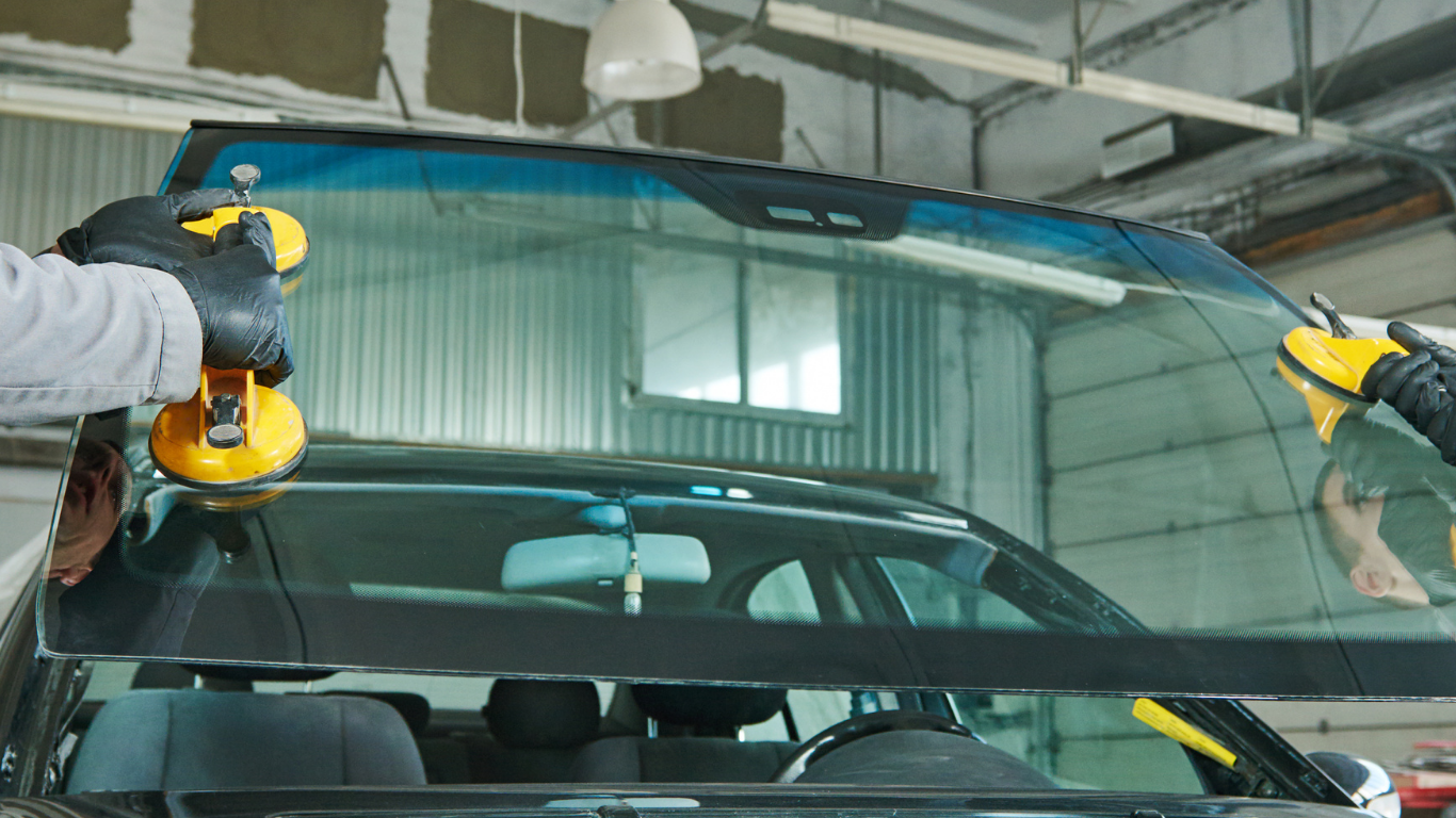 Two workers in gloves use suction tools to remove a windshield in an auto shop