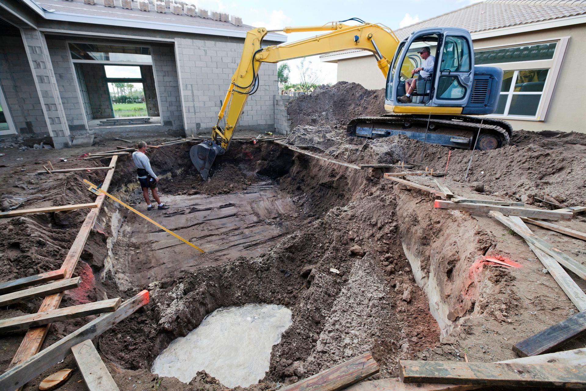 A yellow excavator is digging a hole in the ground in front of a house under construction.