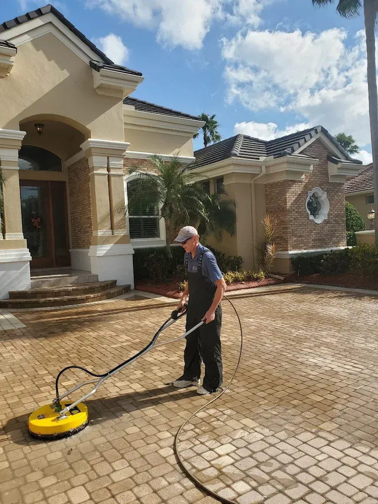 Man pressure washing a brick driveway beside a house with a yellow surface cleaner.