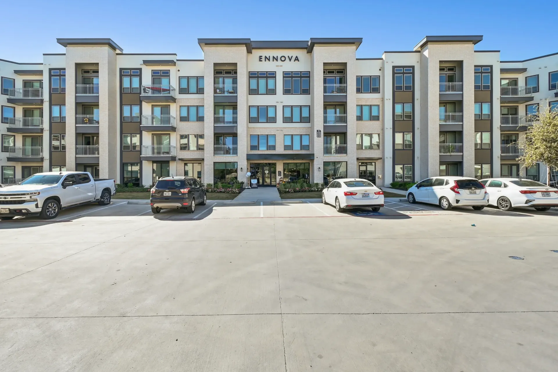 Front of a modern apartment building with balconies and a parking lot in front at Ennova Highlands apartments in Arlington, TX.