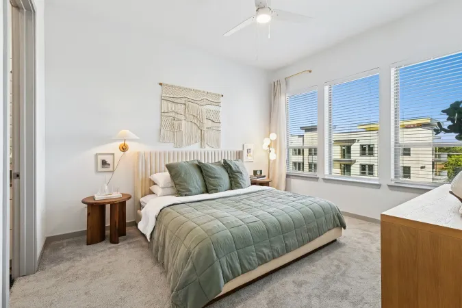 A bright bedroom features a green-quilted bed, a woven wall hanging, a wooden nightstand, and large windows with blinds.