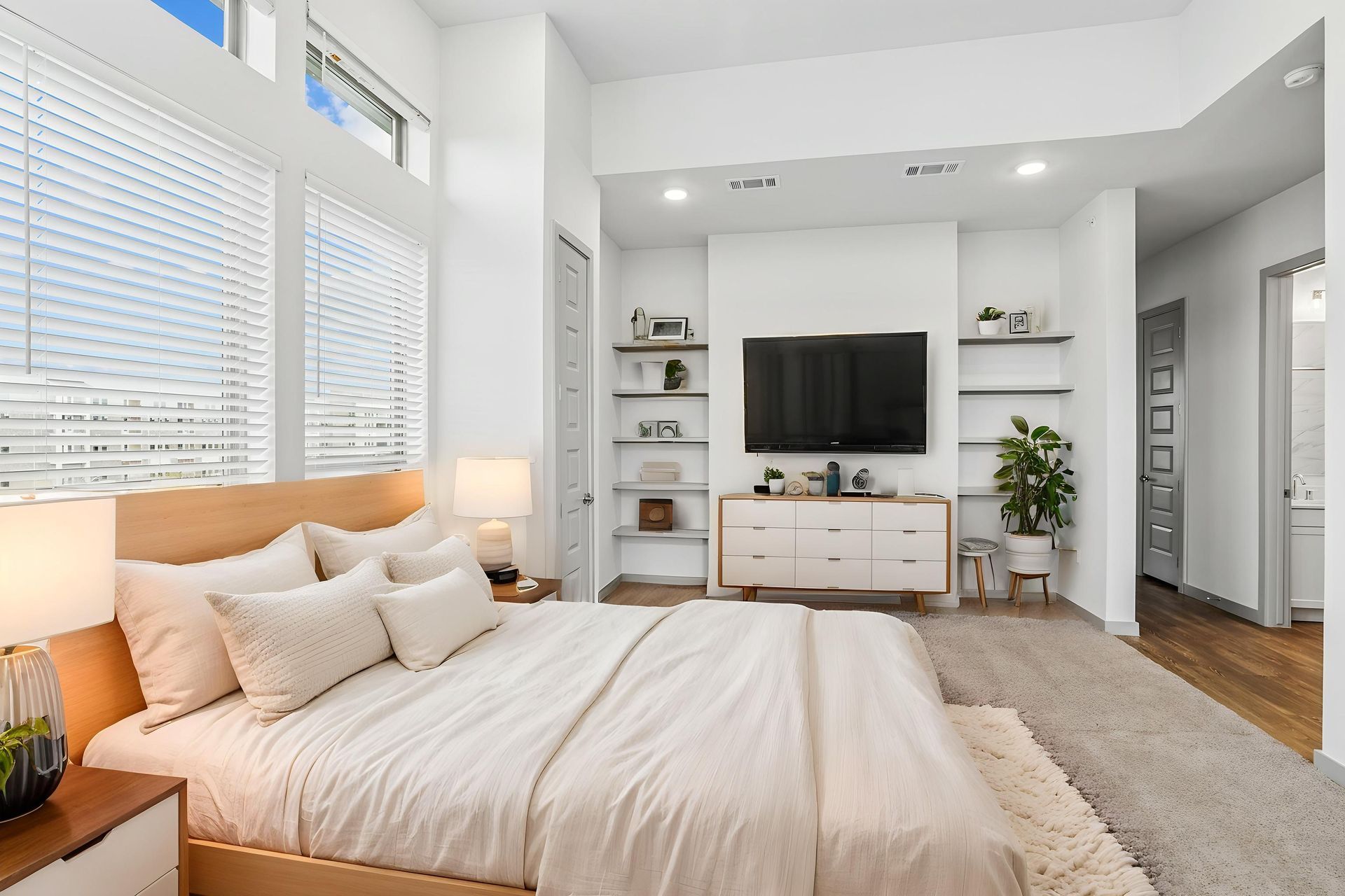 Bright modern bedroom with an unmade white bed, dresser with TV, shelves, and large windows with blinds