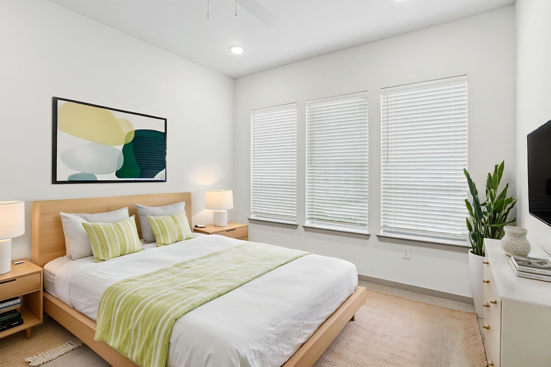 Bright bedroom with a wooden bed, green accents, abstract wall art, and large windows with white blinds
