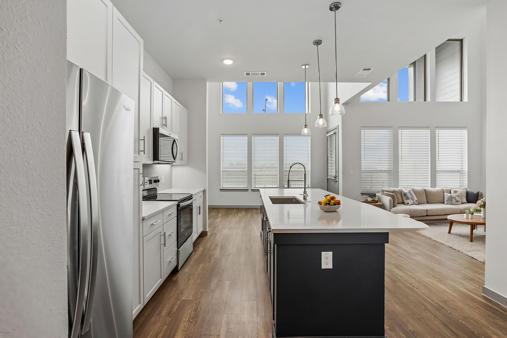Bright modern kitchen with white cabinets, stainless appliances, wood floors, and island open to a living room.
