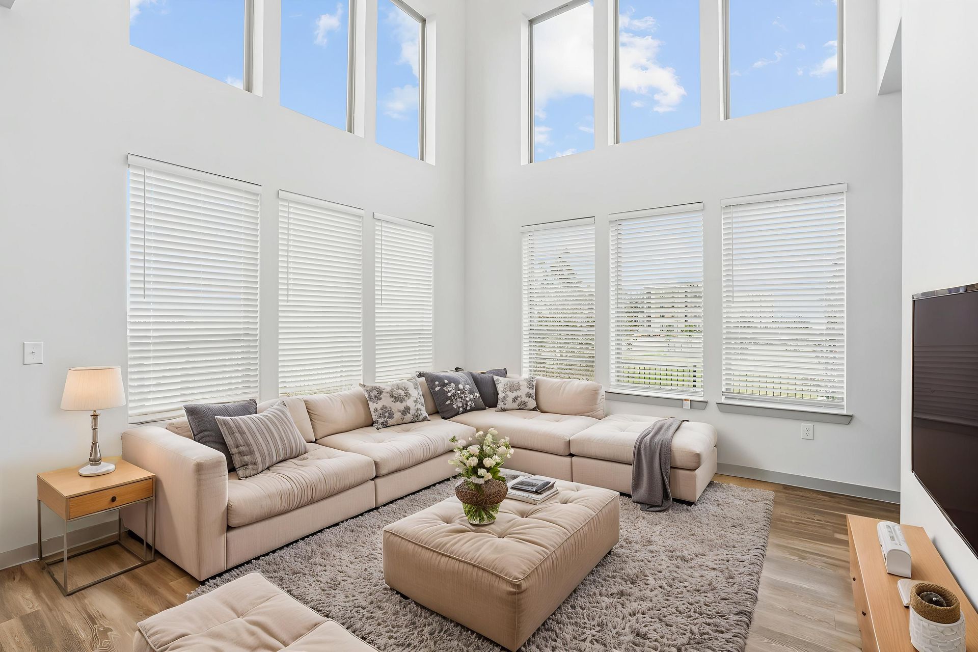 Bright living room with beige sectional, ottoman, gray rug, tall windows, and white shutters