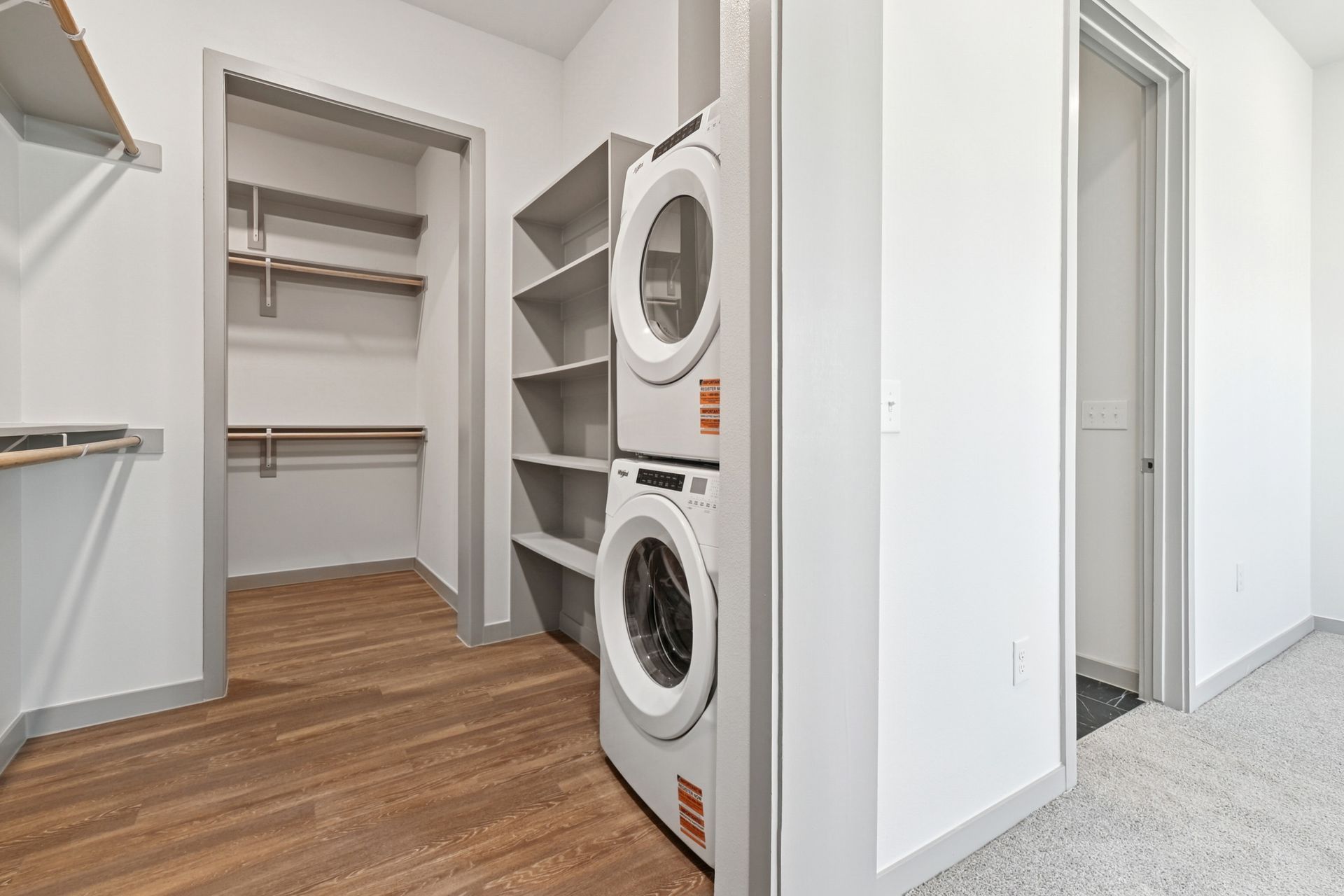 Laundry room with stacked washer and dryer, open shelving, and adjacent hallway with carpet and wood floors
