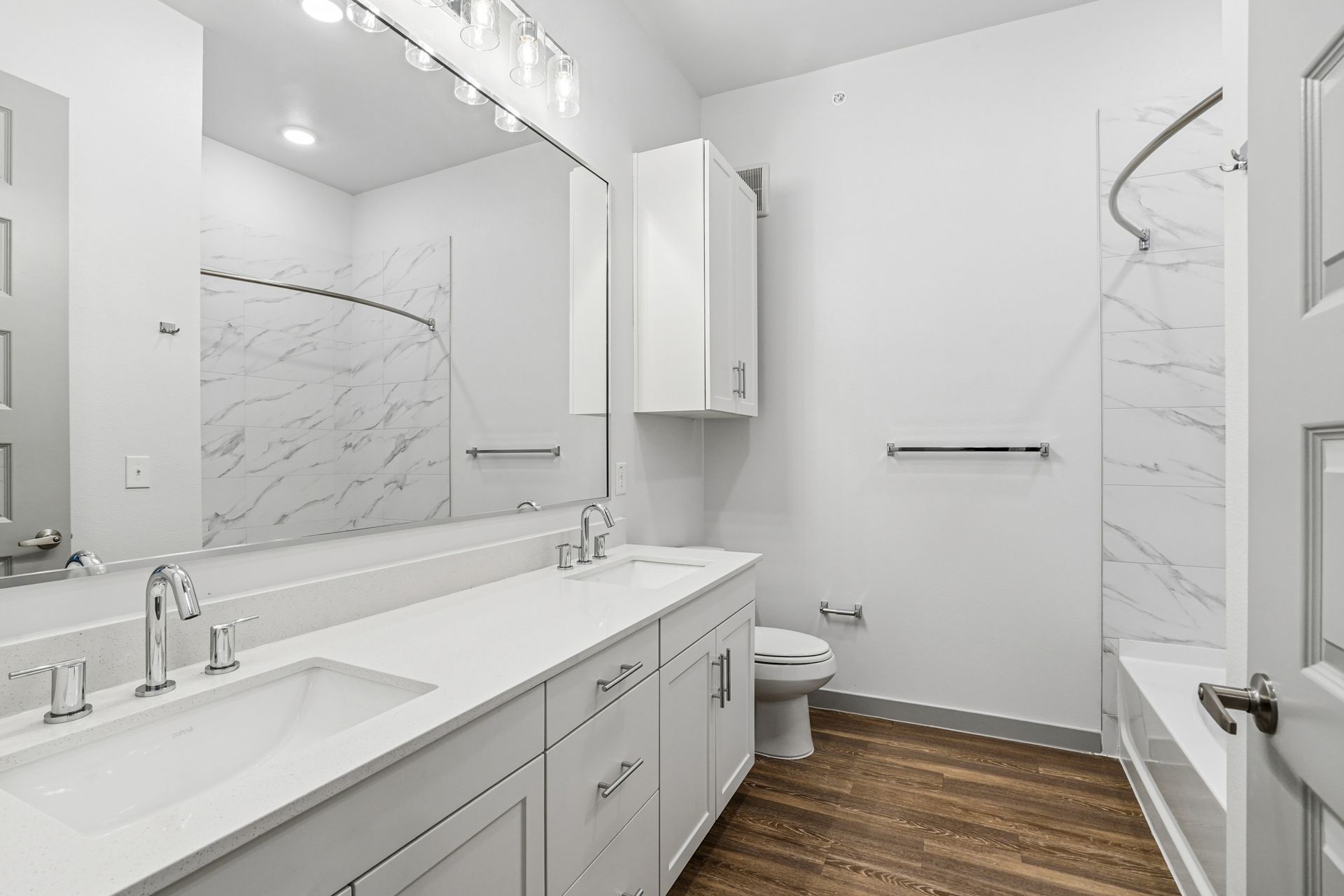 Bright modern bathroom with double vanity, large mirror, tub, toilet, and wood-look floor tile.
