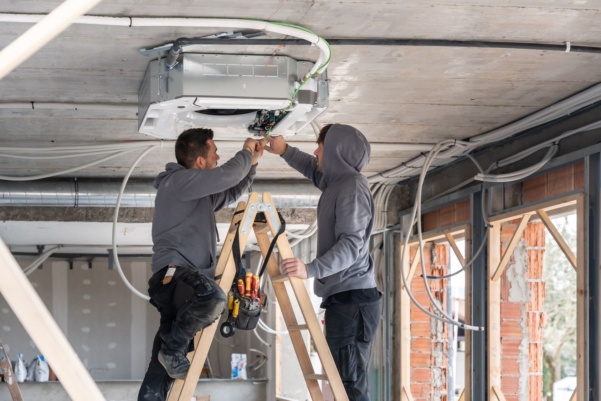 Two workers installing a ceiling-mounted air conditioner in a room under construction.
