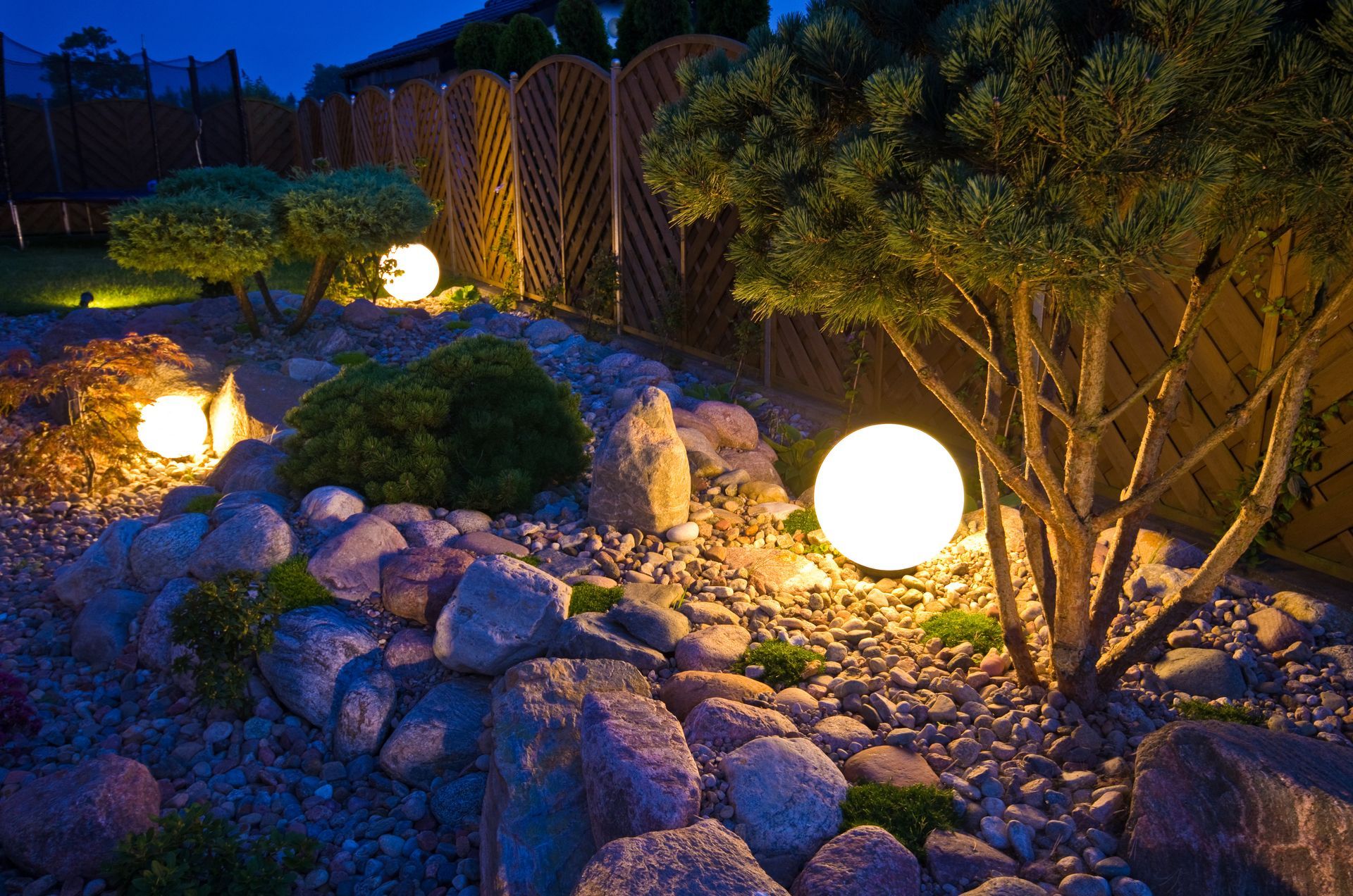 Illuminated garden path with glowing spherical lights, rocks, shrubs, and a wooden fence at dusk Illuminated garden path with glowing spherical lights, rocks, shrubs, and a wooden fence at dusk