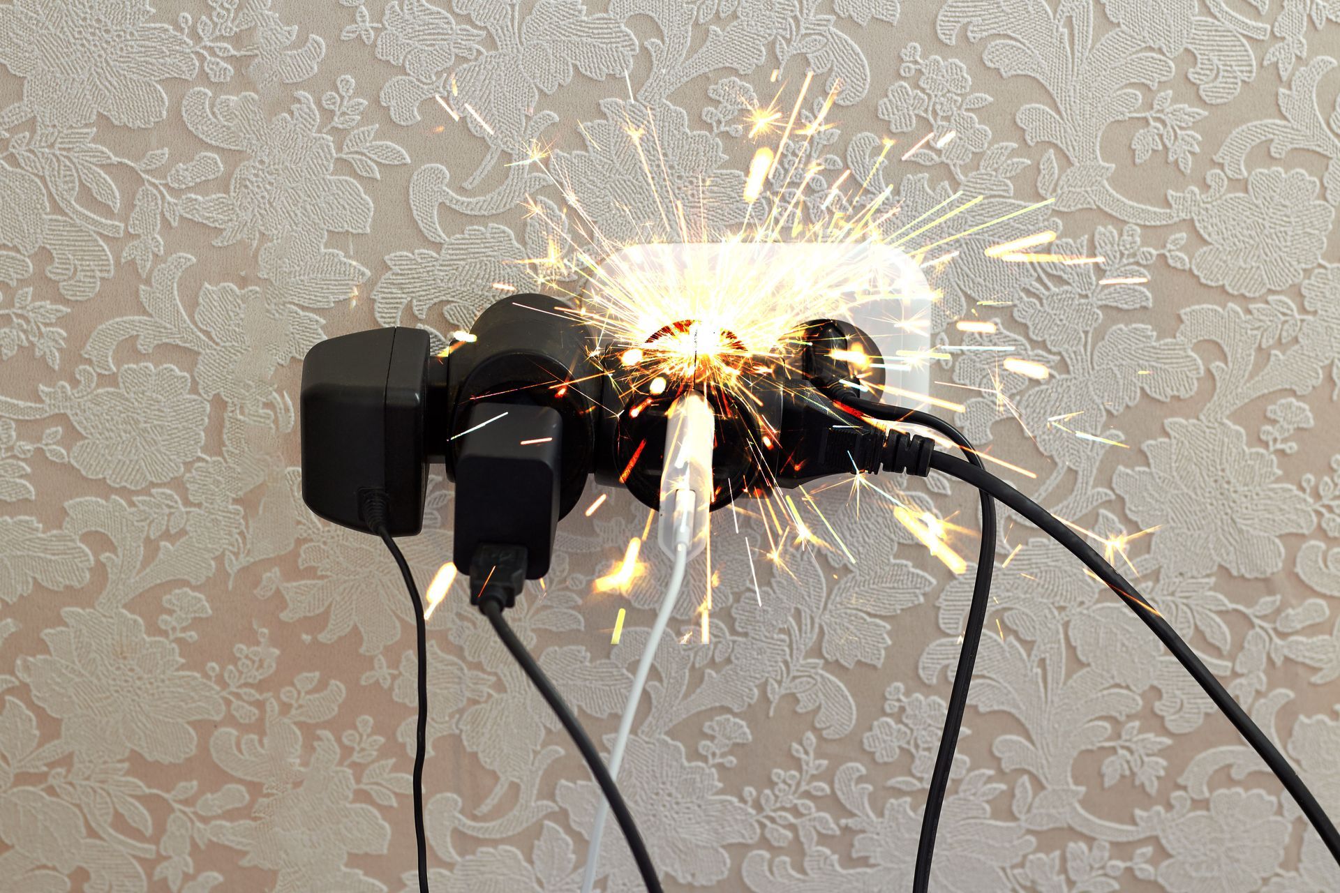 Power strip sparking and catching fire against a patterned wall with several plugged-in cords Power strip sparking and catching fire against a patterned wall with several plugged-in cords