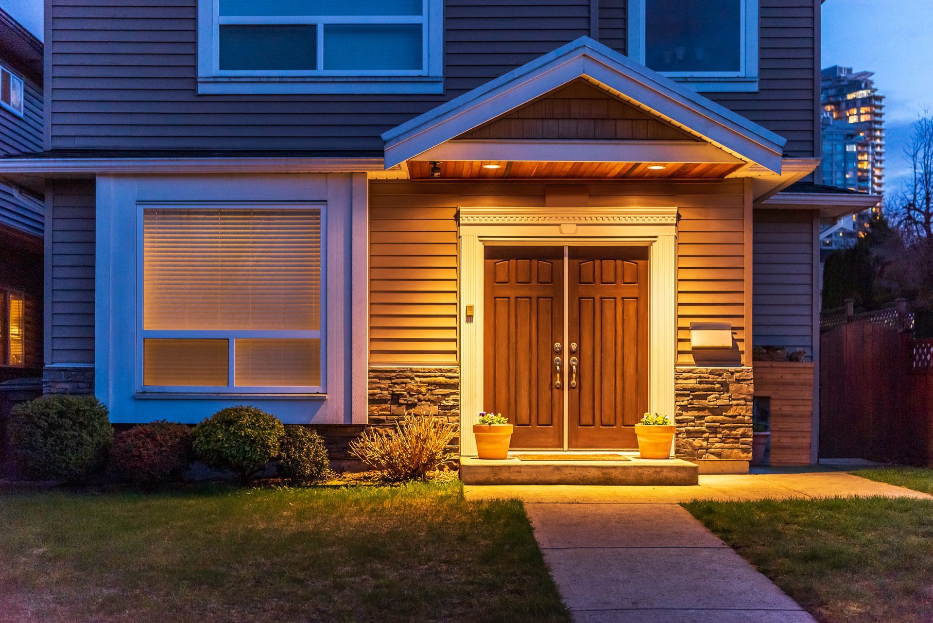 A warmly lit house entryway at dusk with wooden double doors, stone accents, and a small potted plant. A warmly lit house entryway at dusk with wooden double doors, stone accents, and a small potted plant.